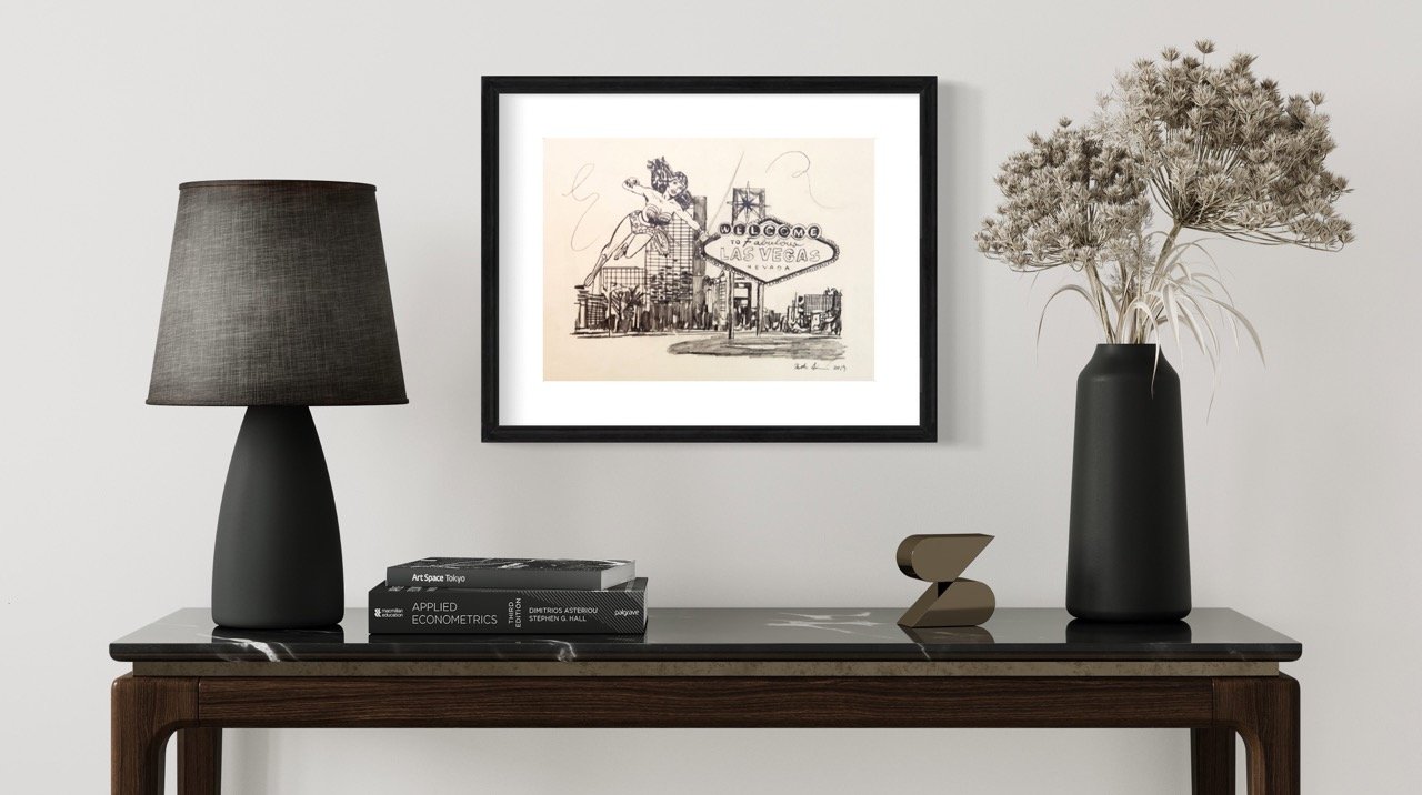 Sketch wonder woman flying next to the "Welcome to Fabulous Las Vegas" sign with energetic scribbles, displayed on a white wall between a textured black lamp and a vase of dried flowers on a console table.