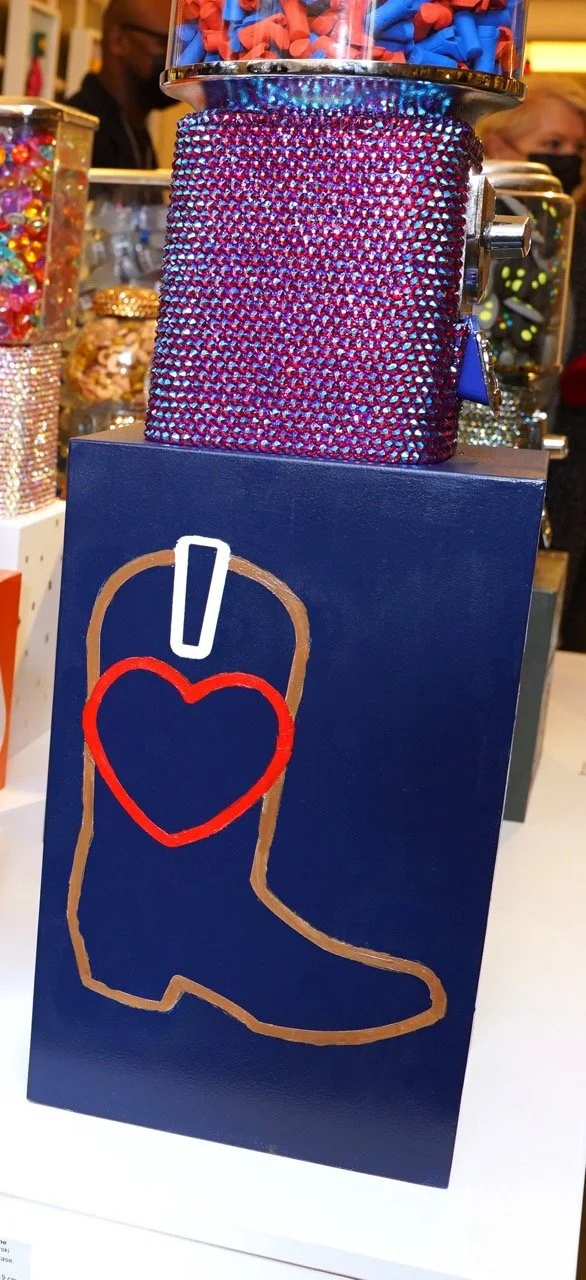 Colorful candy dispenser adorned with rhinestones on top, featuring a blue base with a brown cowboy boot outline and a red heart inside. The background includes various other jars filled with colorful candies.