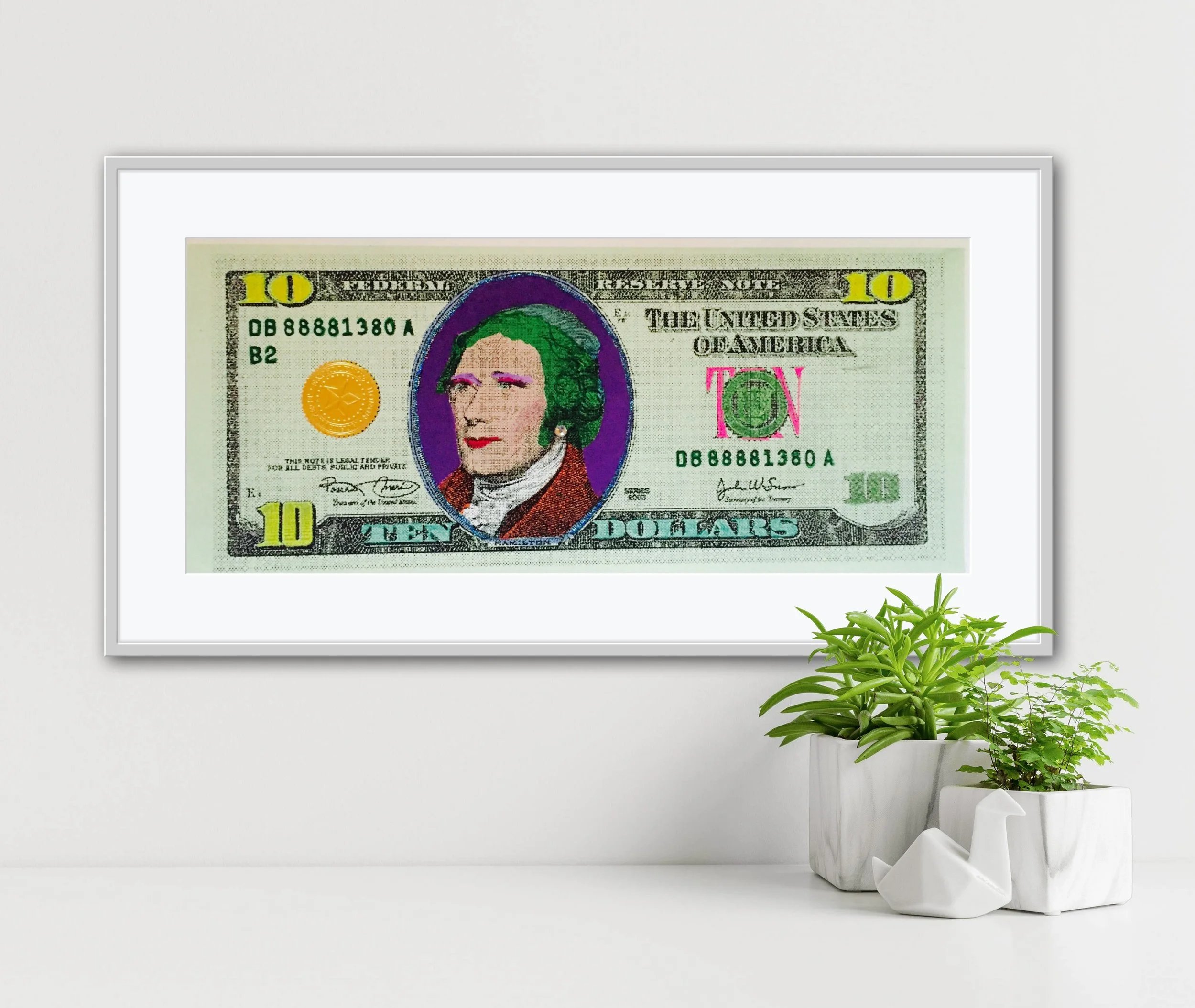 Framed artwork of a modified $10 bill featuring Alexander Hamilton with green hair, purple background, and pink makeup. Set against a clean white wall with two potted plants below, blending pop art with everyday decor.