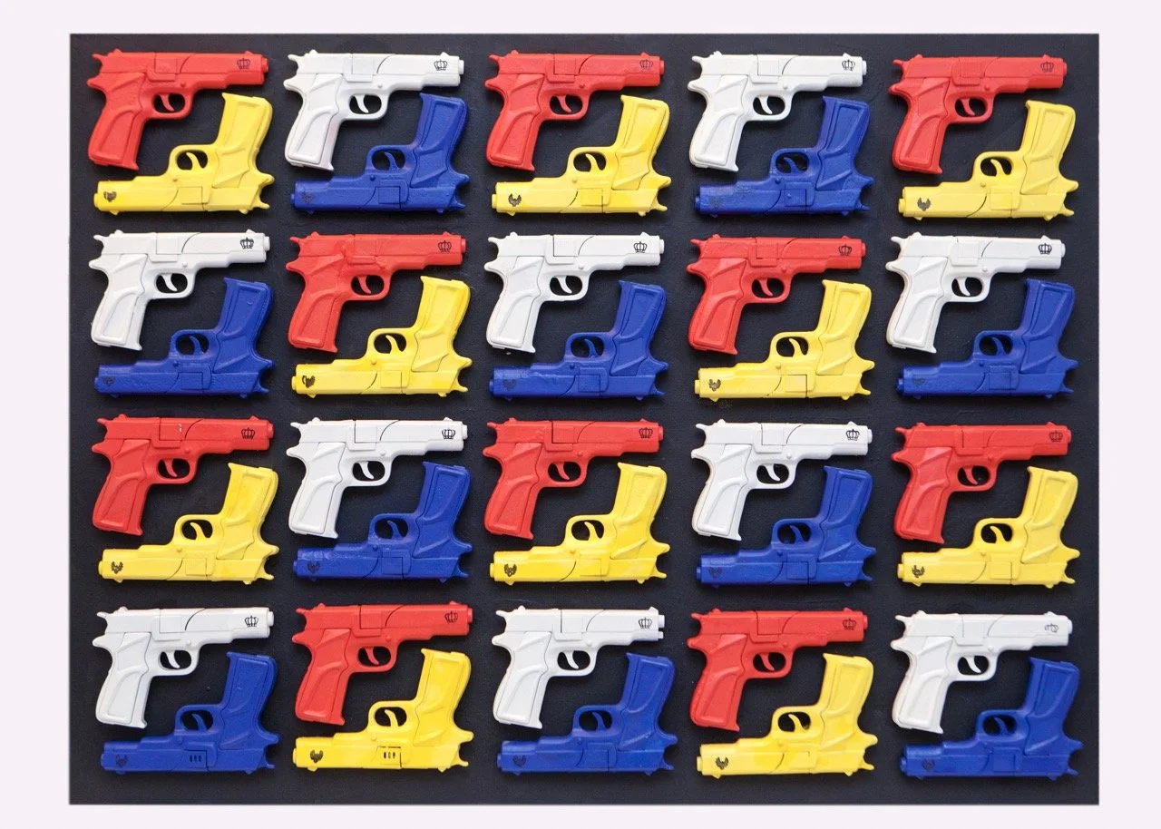 A pop art display featuring a grid of colorful toy guns arranged in rows. The guns are in vibrant shades of red, white, blue, and yellow.