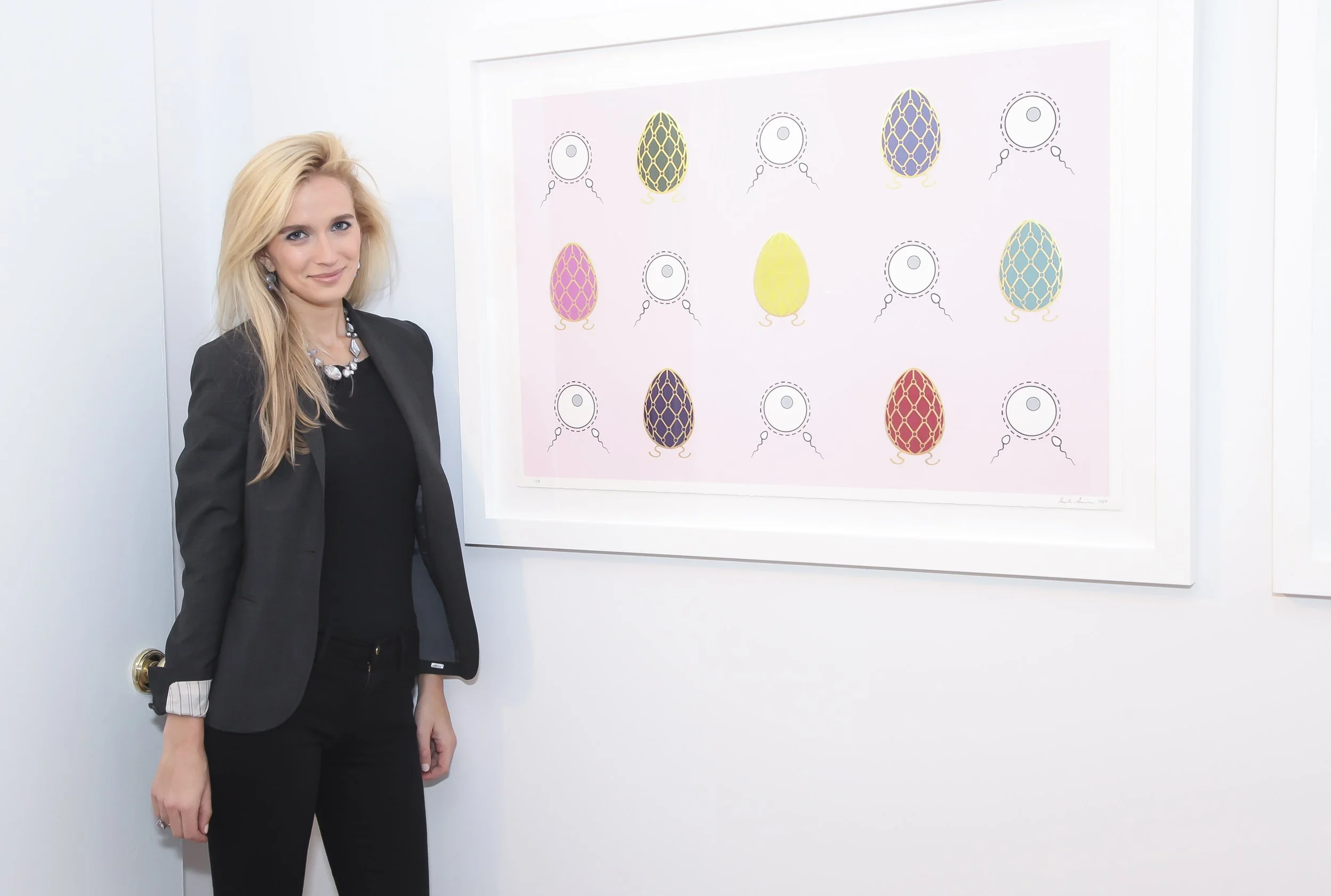 Kristin Simmons stands beside a framed pop art screenprint featuring a grid of colorful Fabergé eggs and sperm fertilizing eggs on a pastel pink background. The Fabergé eggs are in various shades of pink, yellow, green, blue, and purple.