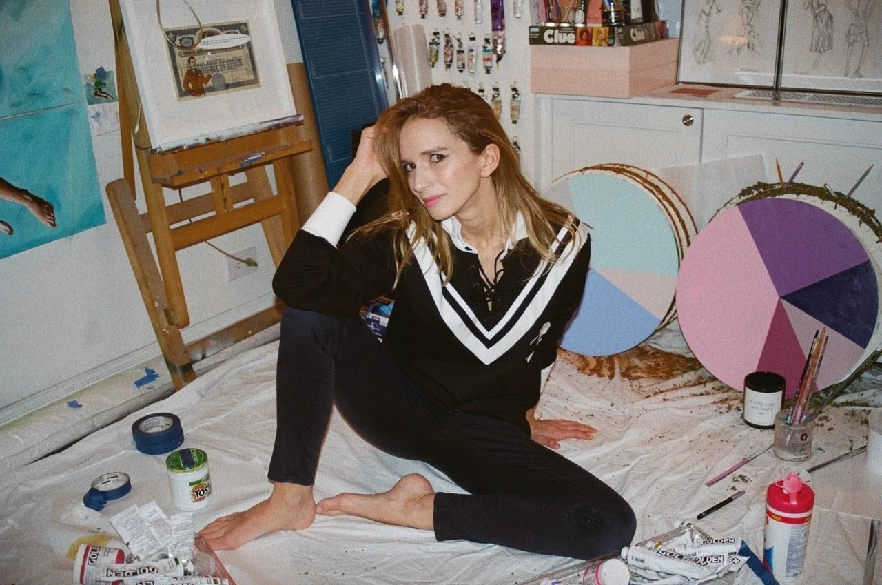 Artist sitting on the floor of a messy studio, surrounded by paint supplies, canvases, and art tools. She is barefoot, wearing a black and white outfit, and resting her head on her hand with a thoughtful expression.