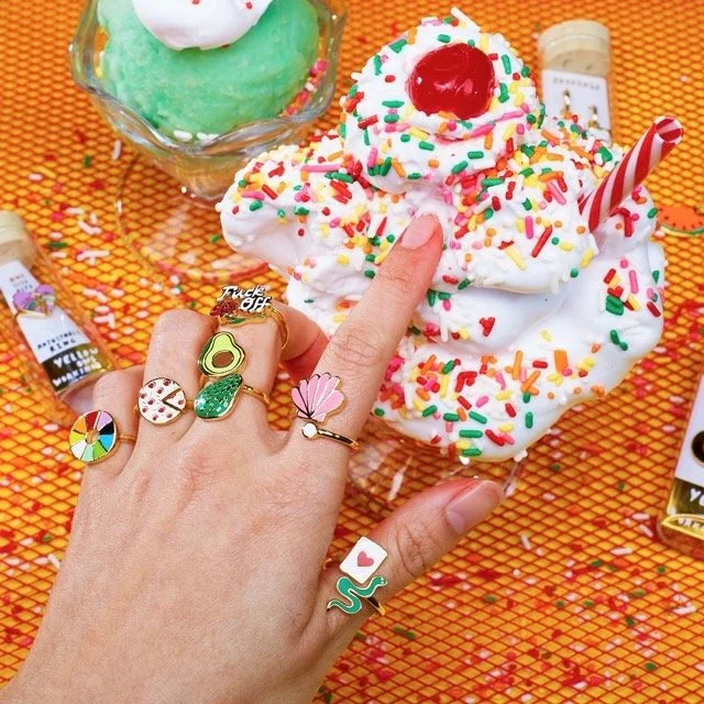 Hand wearing colorful enamel adjustable rings including artist palette, avocado, cactus, shell and snake designs