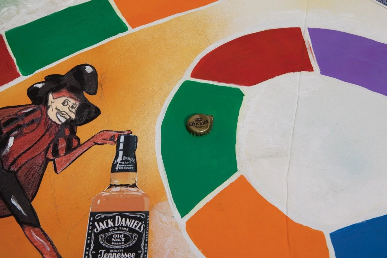 Close up of a reimagined Candy Land board game featuring a whimsical character in a hat holding a bottle of Jack Daniel's whiskey. The background has vibrant sections in green, red, orange, and purple, with a Corona beer bottle cap placed near the ce