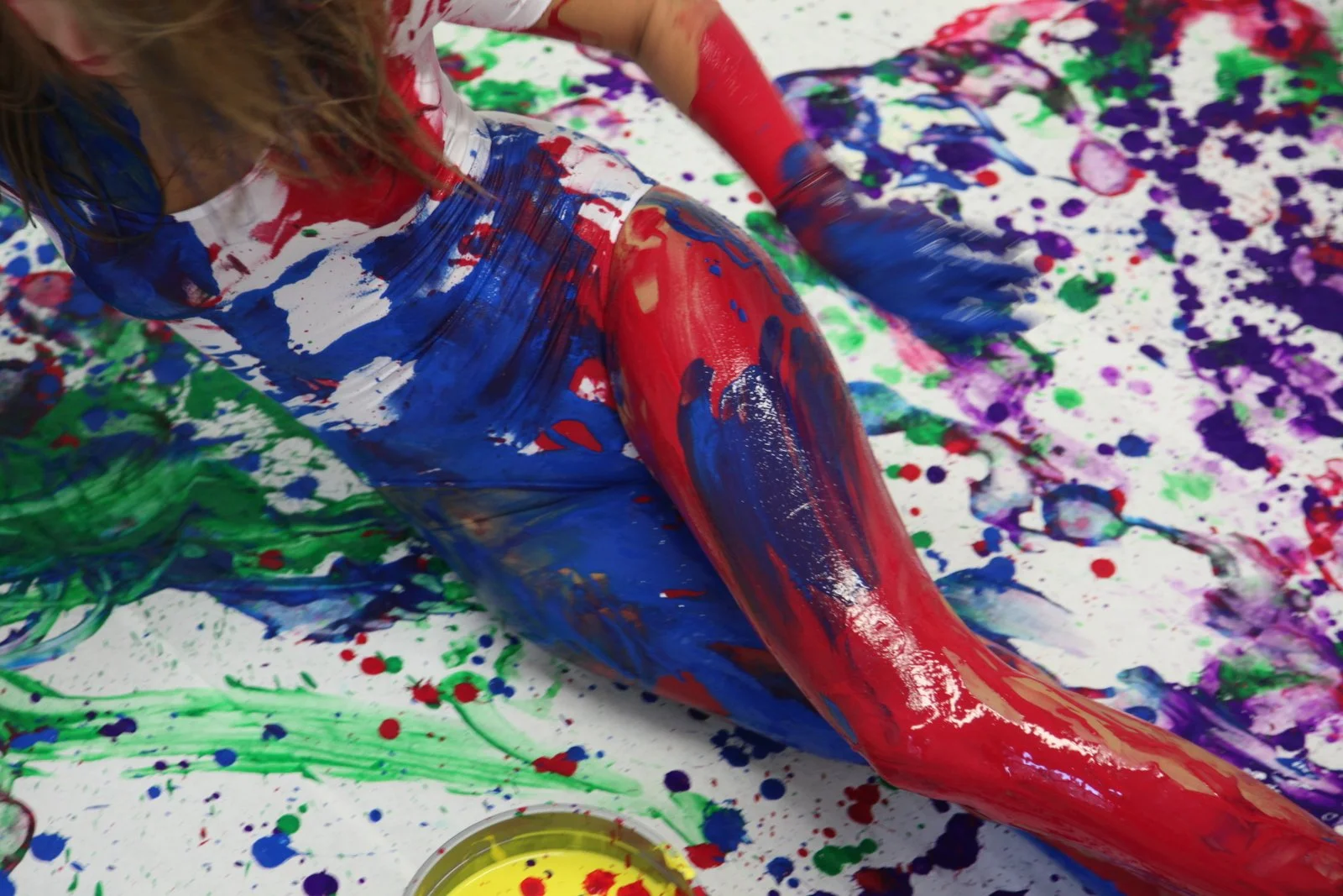 Body painted in red, blue, and white, lying amidst a floor covered in green, purple, and blue paint splatters.