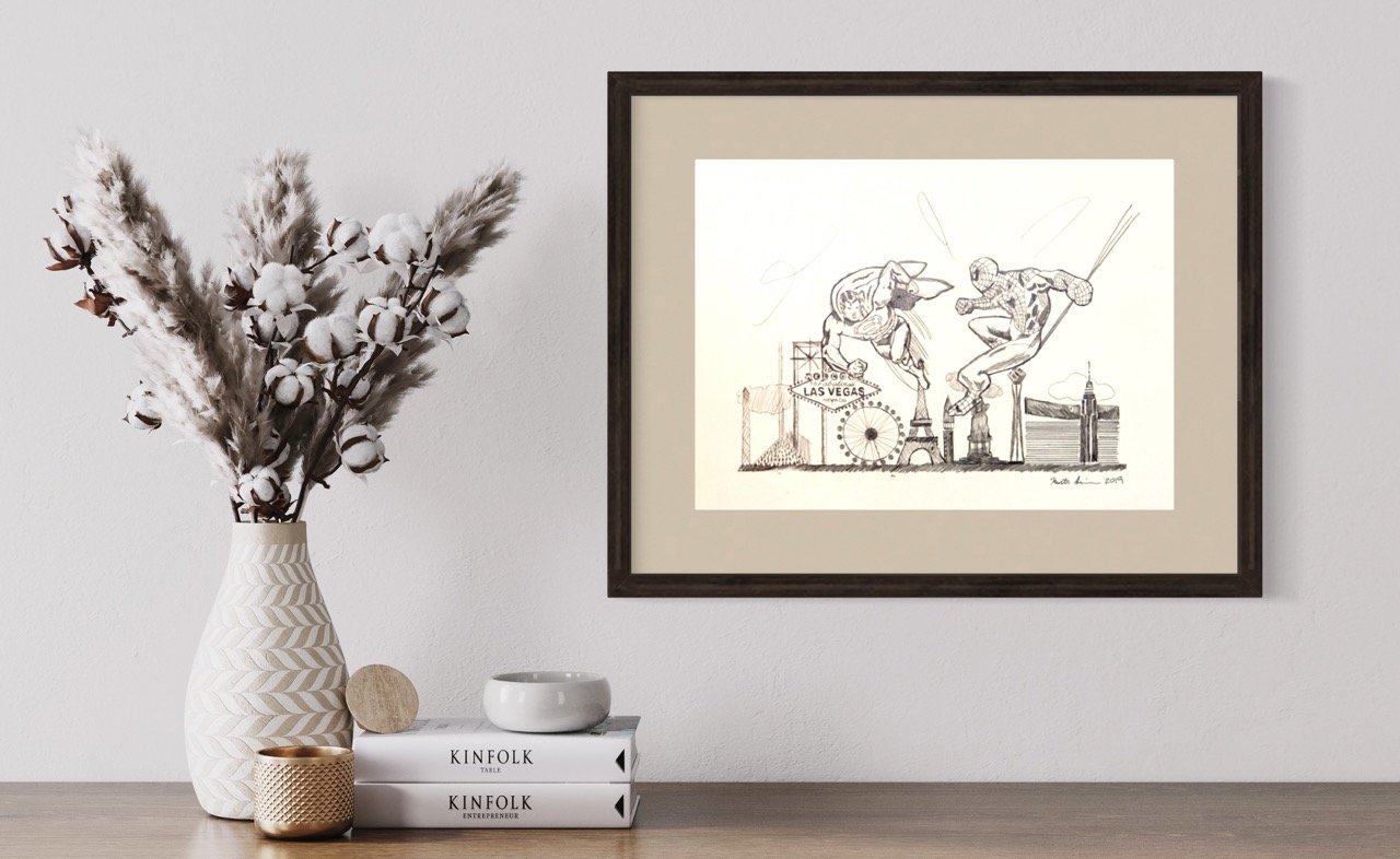 Framed pencil drawing of spiderman and superman in dynamic poses with landmark Las Vegas buildings, set on a wall next to a textured vase filled with cotton branches, atop a surface with stacked books and a small candle.