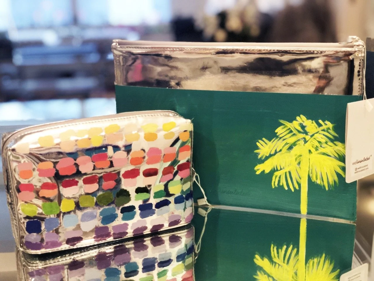 Display of two stylish pouches on a reflective surface. The smaller pouch features a vibrant, abstract polka dot pattern with multicolored spots, while the larger pouch has a sleek, silver top and a green base with a bright yellow palm tree illustrat