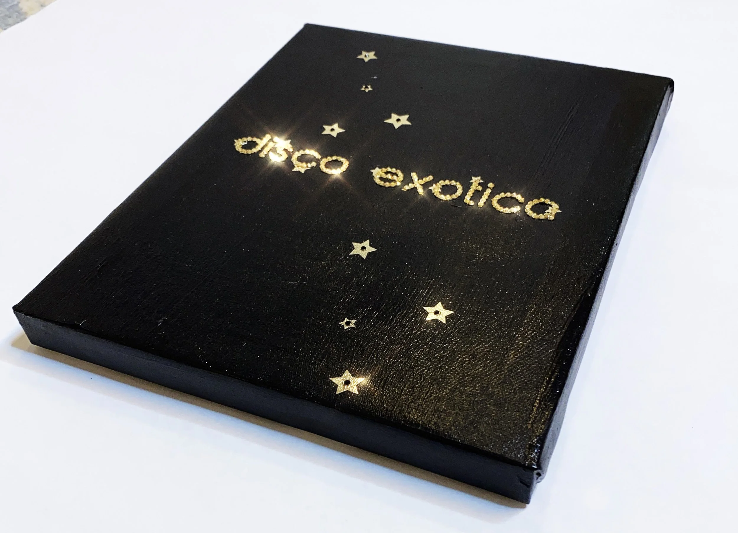 Small black canvas with golden text reading "disco exotica" surrounded by sparkling star-shaped decorations. The shiny gold elements create a glamorous, festive feel against the dark background.