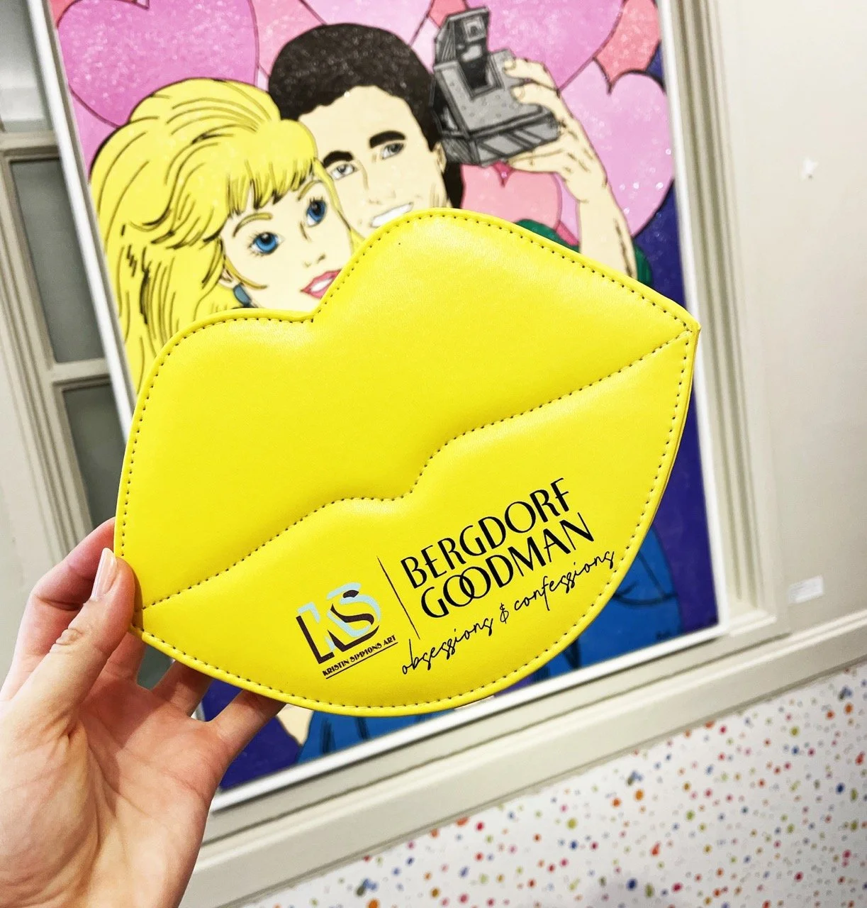 A person holds a bright yellow, lips-shaped clutch with the "Bergdorf Goodman" logo and the text "obsessings & confessings." In the background, a pop art painting depicts Barbie and a dark-haired man taking a selfie