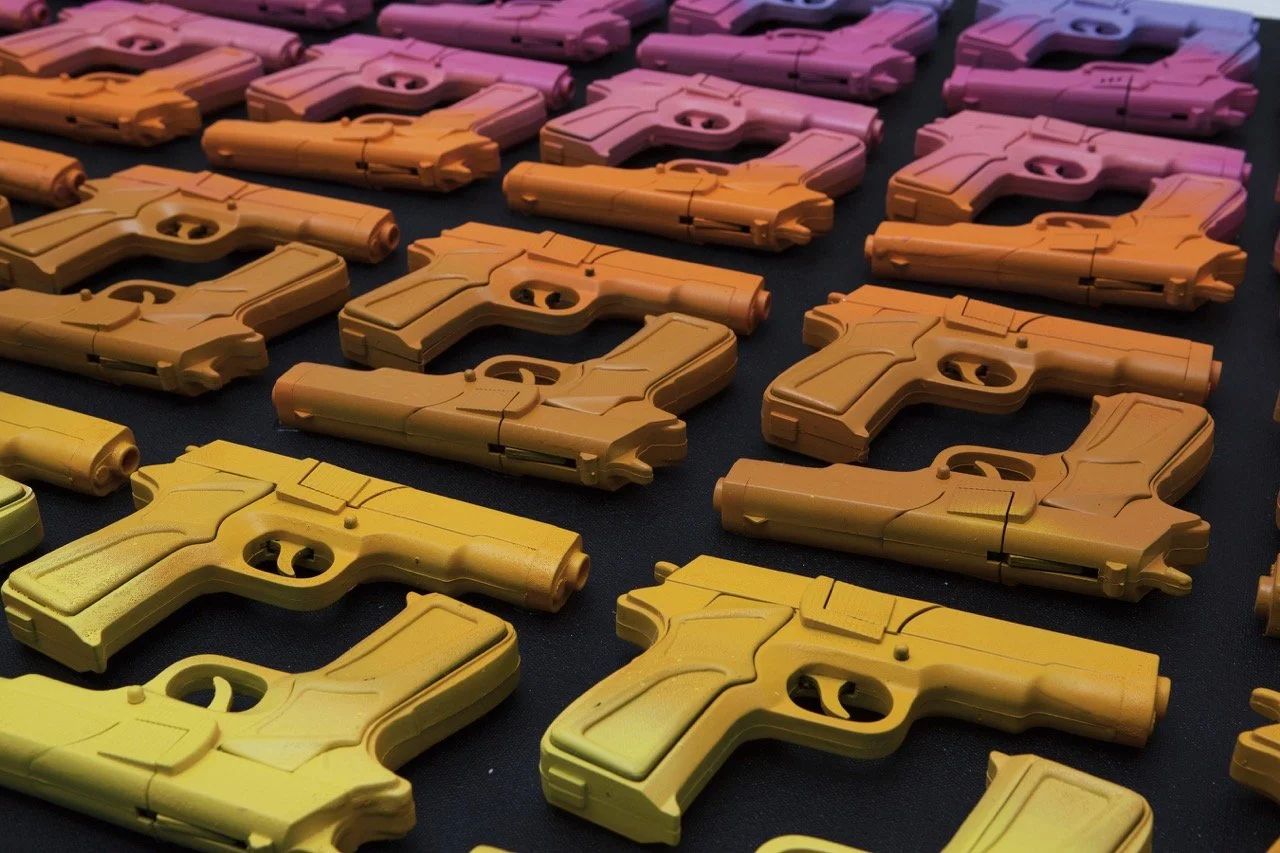 Rows of toy guns in various colors from yellow to purple arranged on a black surface. The guns create a gradient effect, with each row transitioning from one color to the next.
