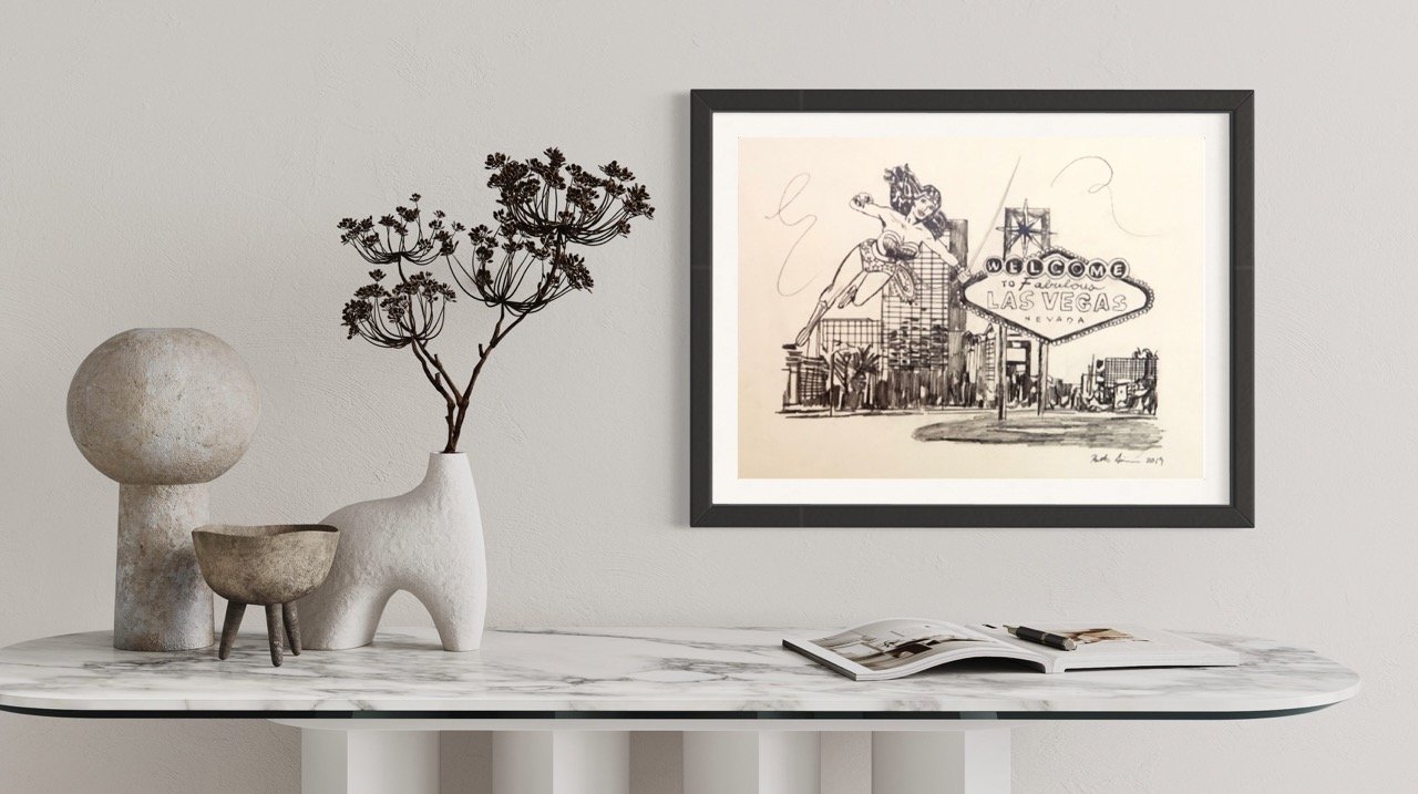 "Pencil drawing of Wonder Woman comic character next to the Las Vegas sign in a frame on a wall above a marble table with abstract ceramic sculptures and a dried floral arrangement."