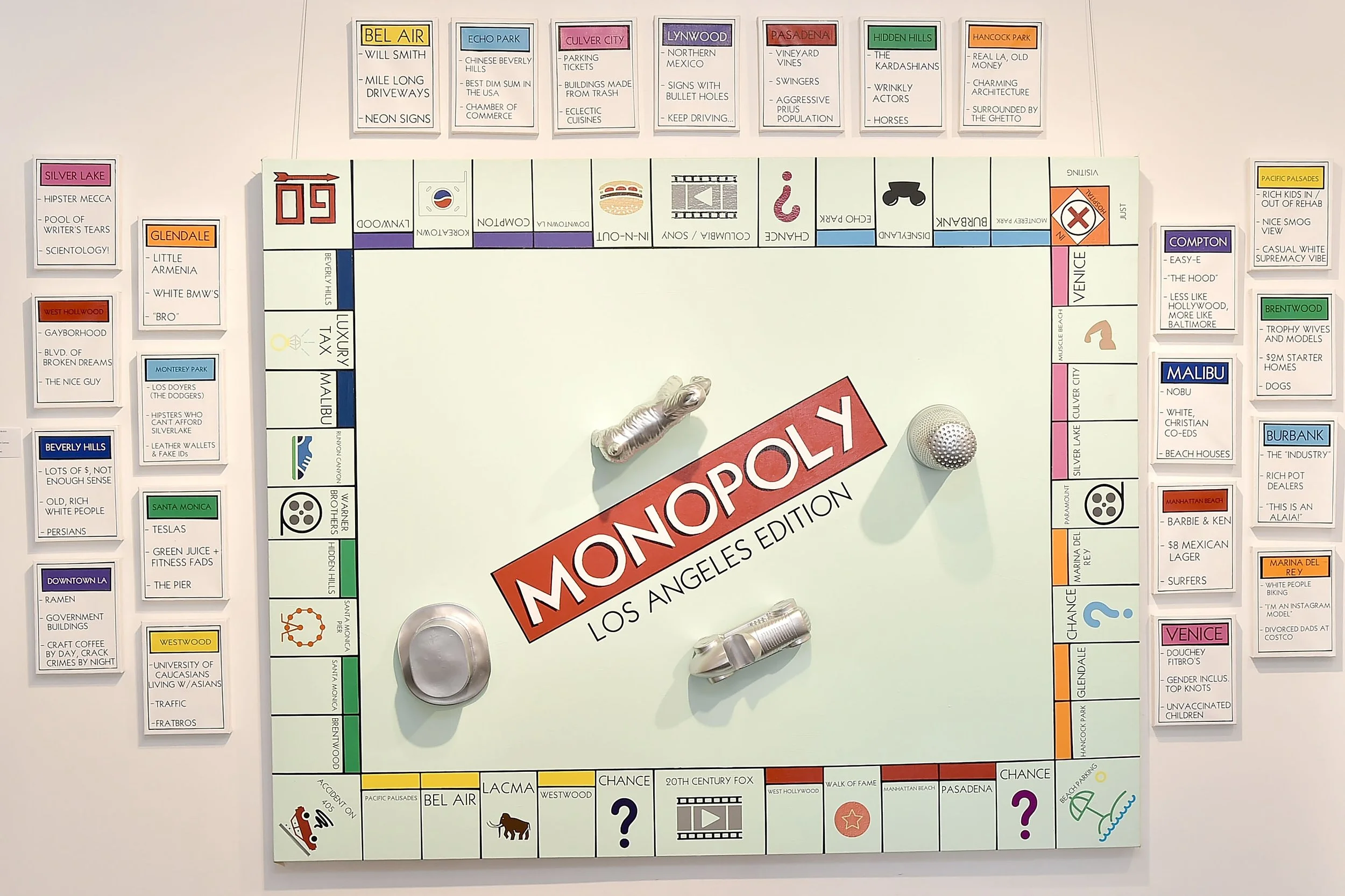 Monopoly board titled "Los Angeles Edition" featuring properties with humorous descriptions of LA neighborhoods. The center of the board displays "Monopoly" in red, surrounded by various colored property spaces and oversized game tokens, including a 