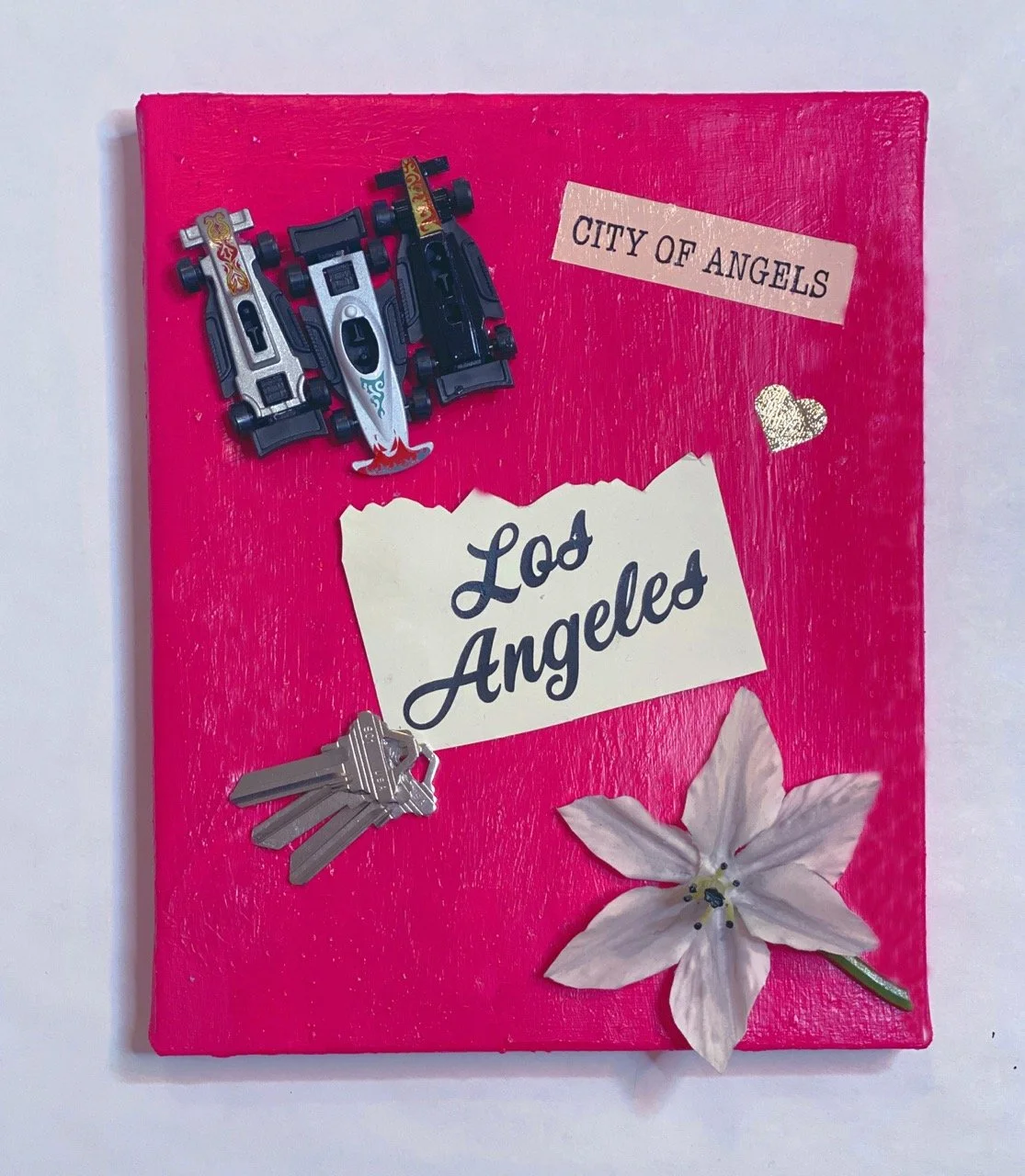Vibrant mixed-media artwork on a hot pink background featuring miniature race cars, a "City of Angels" label, a note reading "Los Angeles," a set of keys, and a fabric flower.