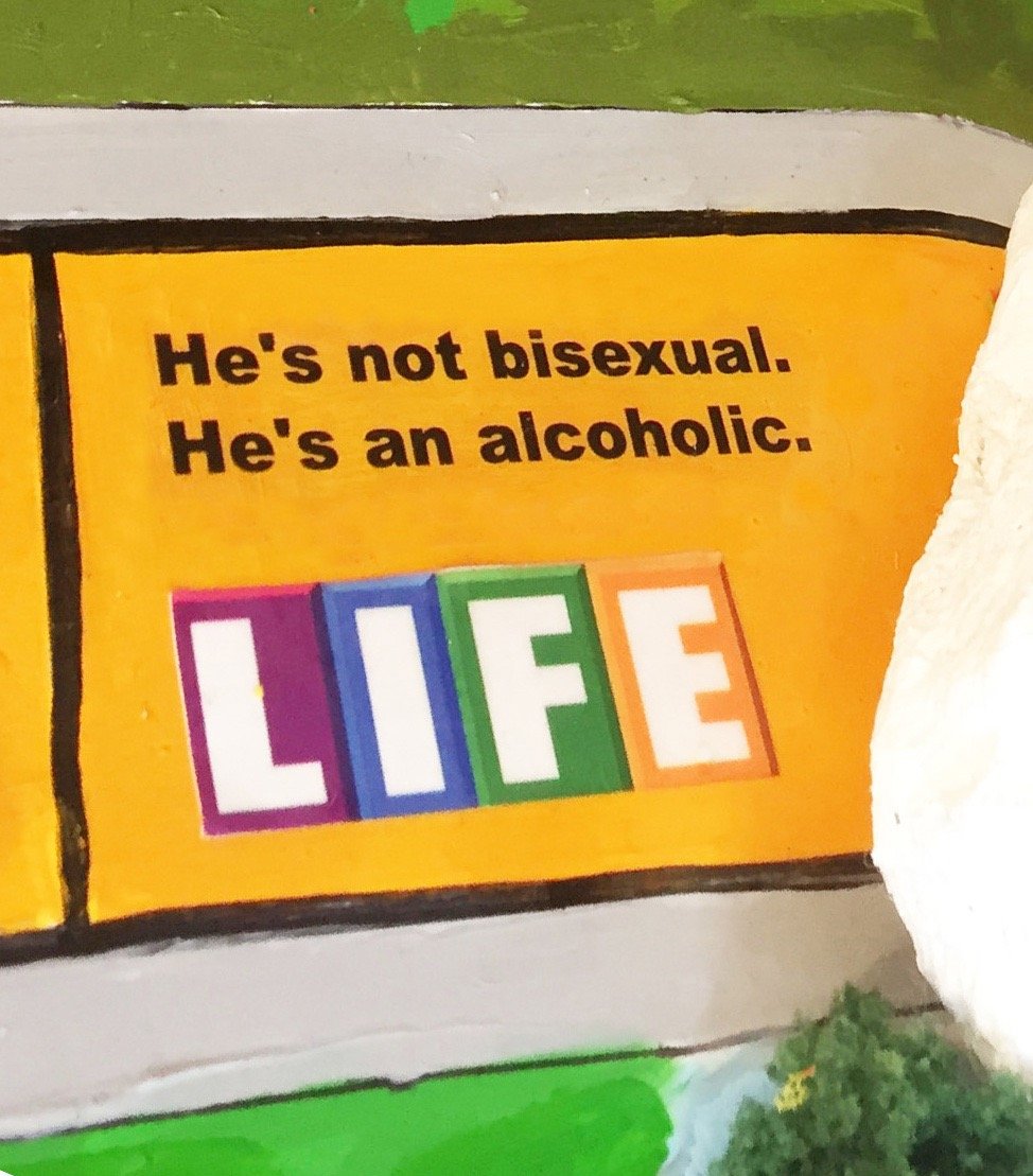 Brightly painted Game of LIFE tile with an orange background and the text, "He's not bisexual. He's an alcoholic."