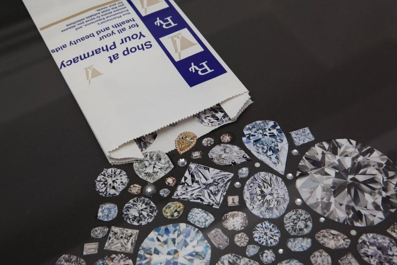 Close-up of a collection of sparkling diamonds in various cuts and colors arranged on a glossy black surface. A white paper bag with blue text, promoting a pharmacy, is partially visible, adding contrast to the dazzling gems.