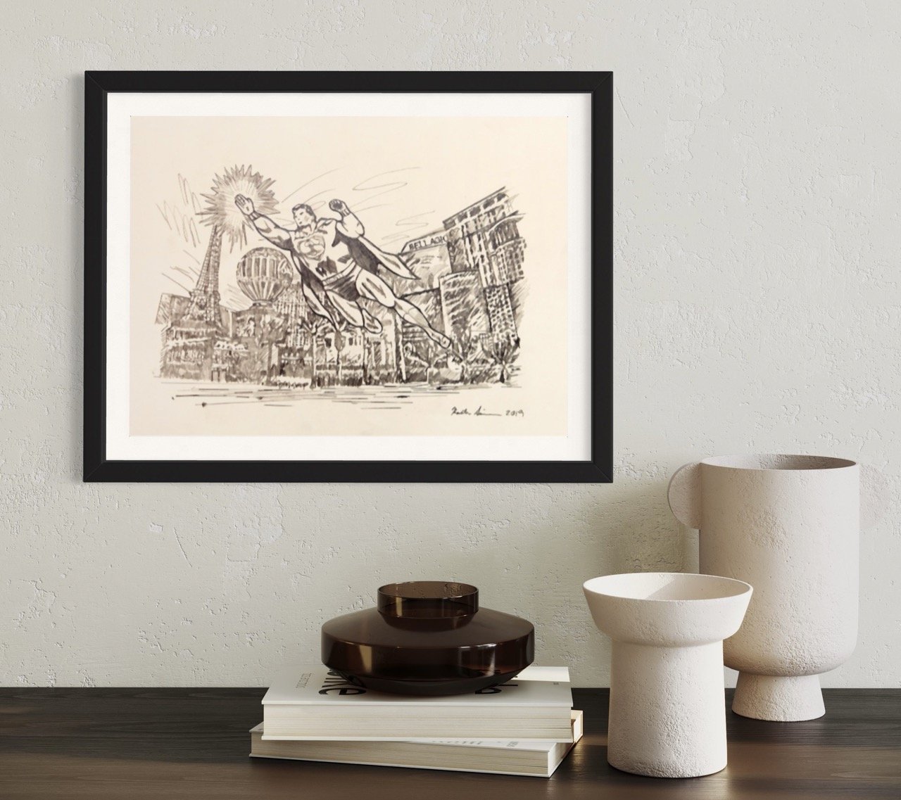 The artwork, framed on a wall, portrays superman in flight against the Las Vegas skyline as a backdrop. Accompanying the scene are modern vases and stacked books on a dark surface.