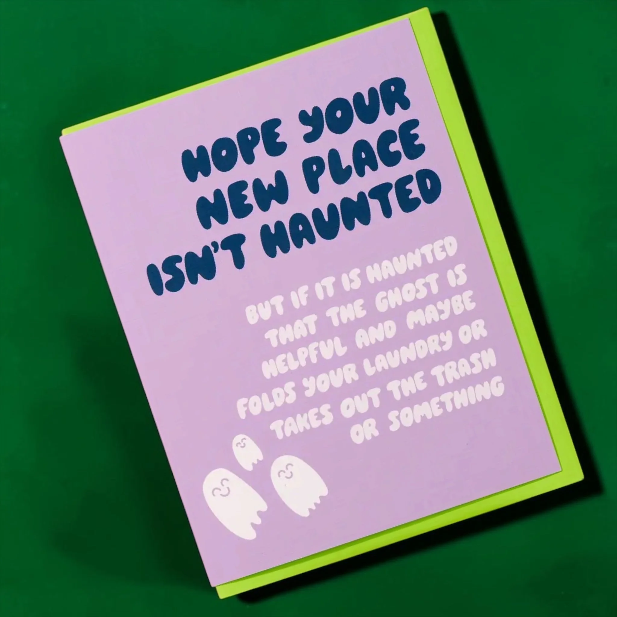 Funny new home card reading “Hope your new place isn’t haunted” on a lavender background with small ghost illustrations and a bright green envelope