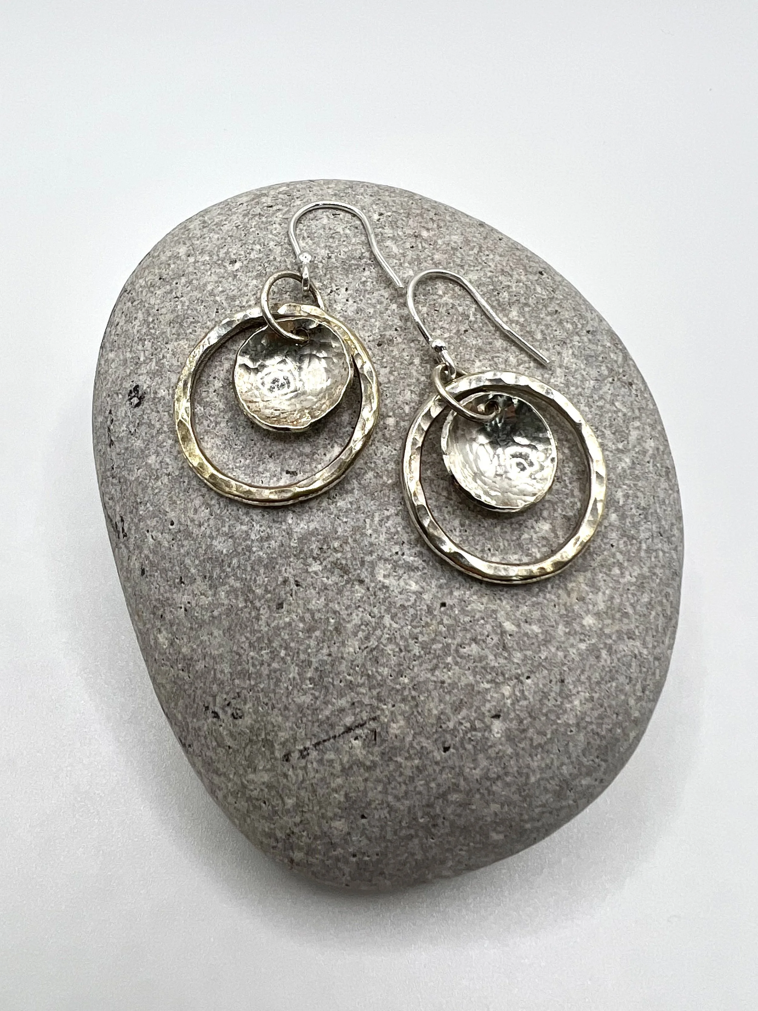 Silver earrings with hammered texture, dangling from hooks, placed on a smooth, oval gray stone.
