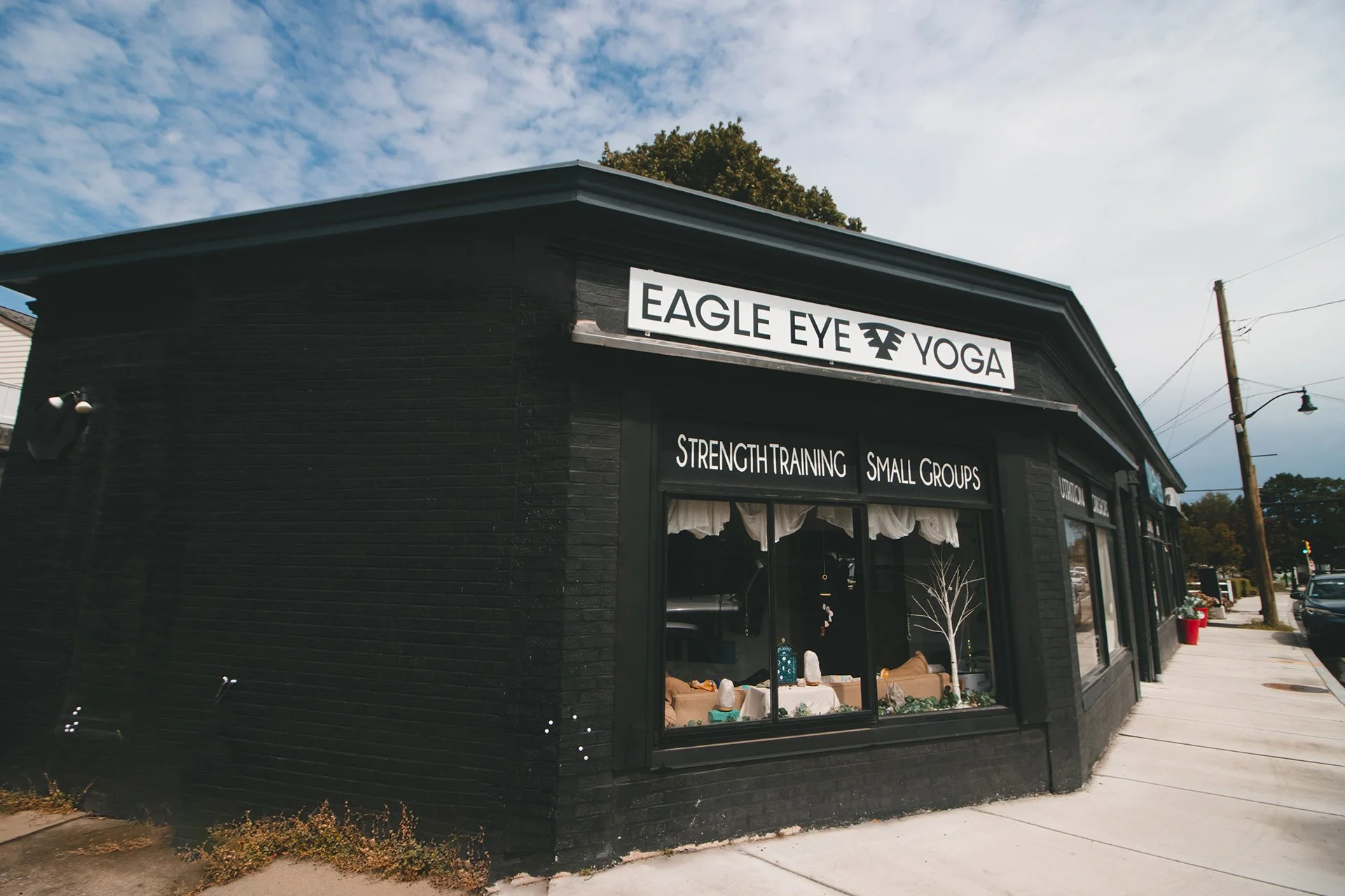 Exterior view of a black building with signs for 'Eagle Eye Yoga' and 'Strength Training Small Groups,' with large windows displaying decor and activities inside, sidewalk in front, and a street with cars and utility poles in the background.