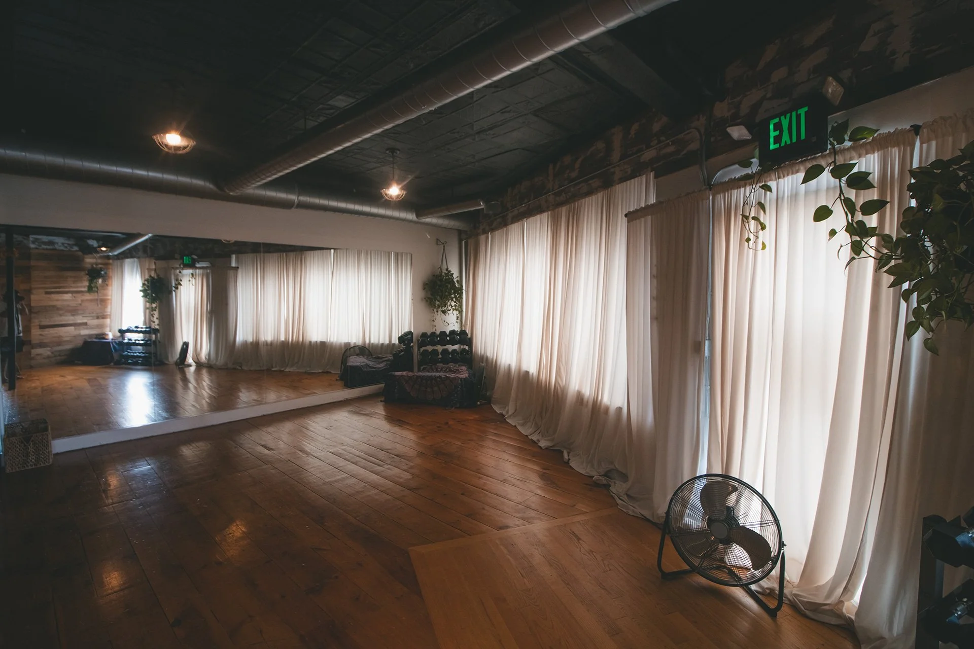 Empty dance studio with wooden floors, large mirrors, beige curtains, potted plants, a fan, and a diamond-shaped rack of weights, illuminated by ceiling lights and natural light.