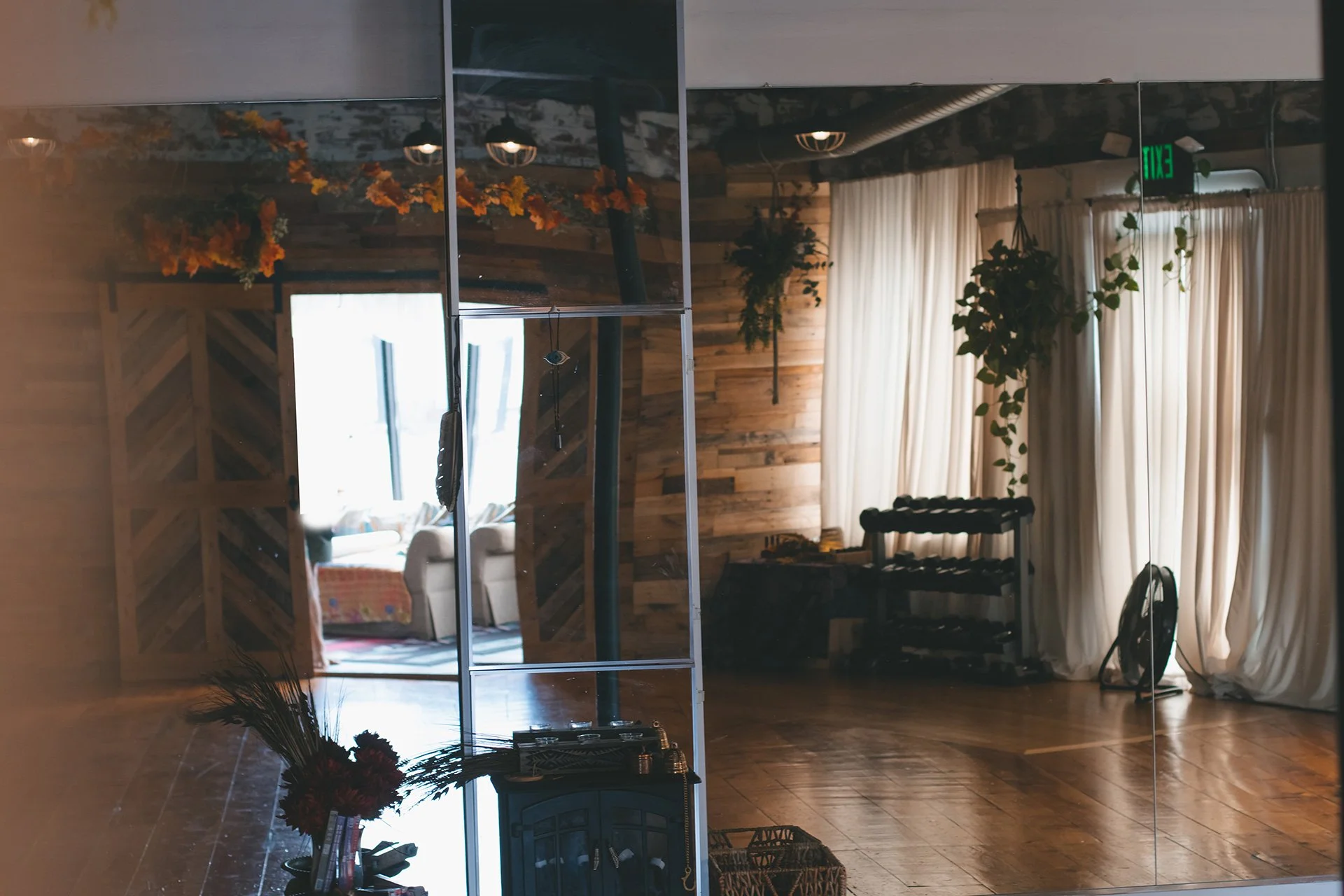 Interior of a room with wooden walls, a large mirror, cream curtains, potted plants, and furniture, reflecting natural light from windows.