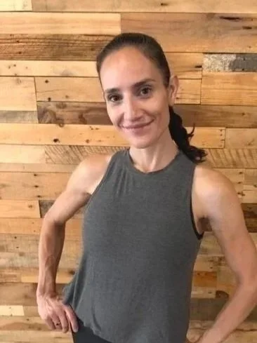 A woman with long, wavy brown hair smiling at the camera, wearing a sleeveless black top, standing in front of a wooden wall.