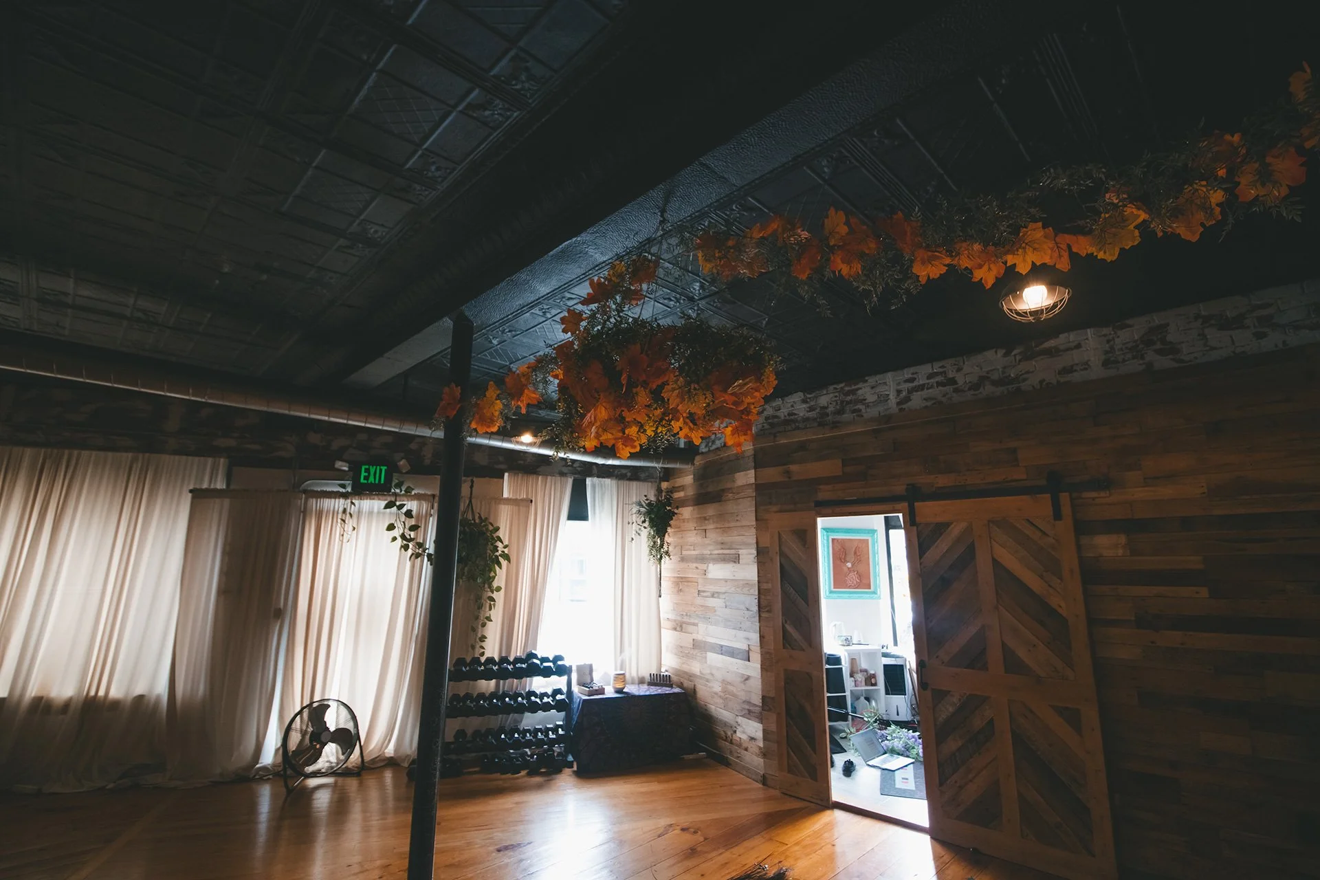 An indoor room with wooden walls, ceiling, and floor. There are curtains, a small fan, wine rack, and an open sliding barn door. Orange flowers and green foliage are hanging from the ceiling.