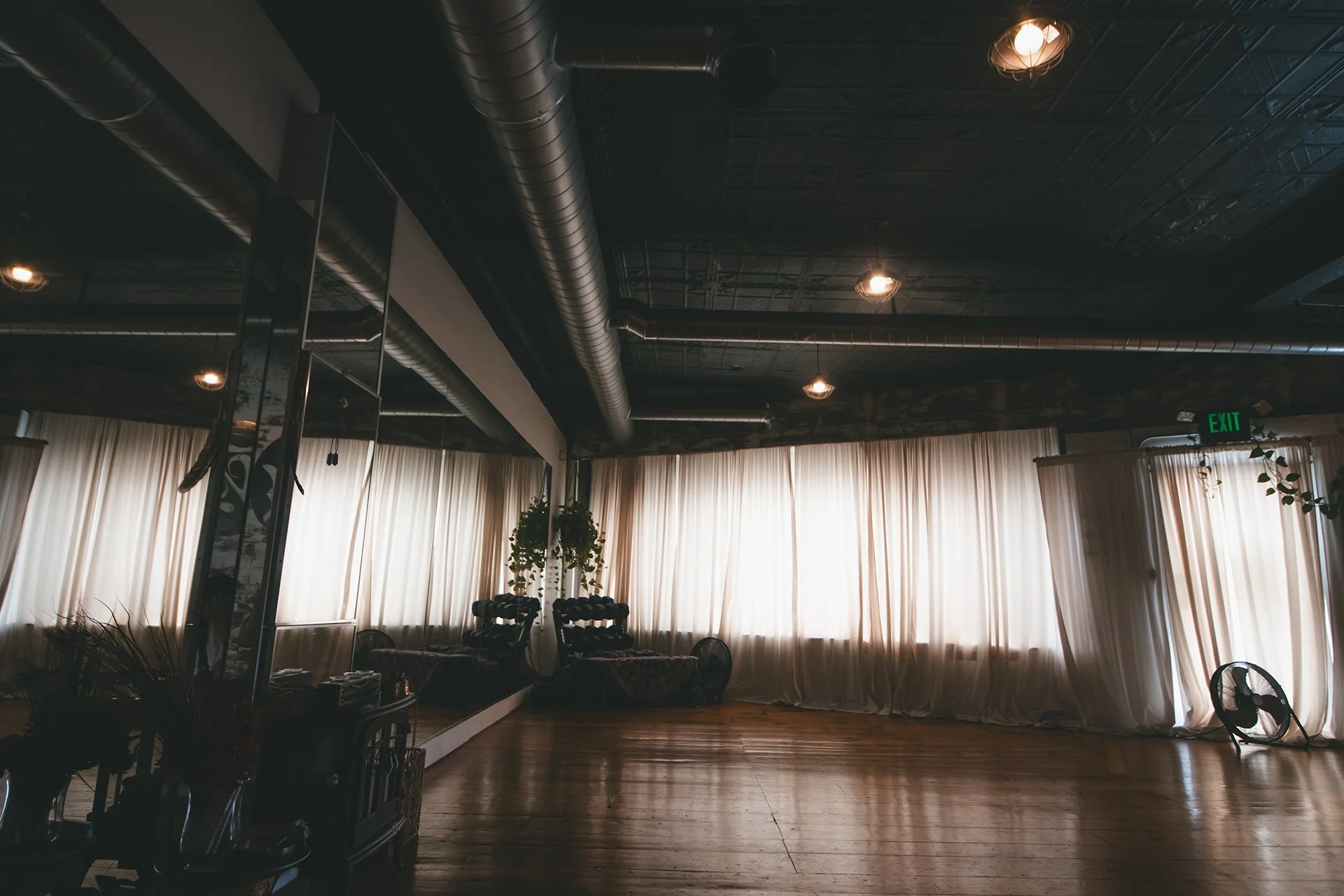 Empty room with wooden floors, large windows with light-colored curtains, potted plants, a black standing fan, ceiling lights, exposed ducts, and a mirror reflecting some furniture.