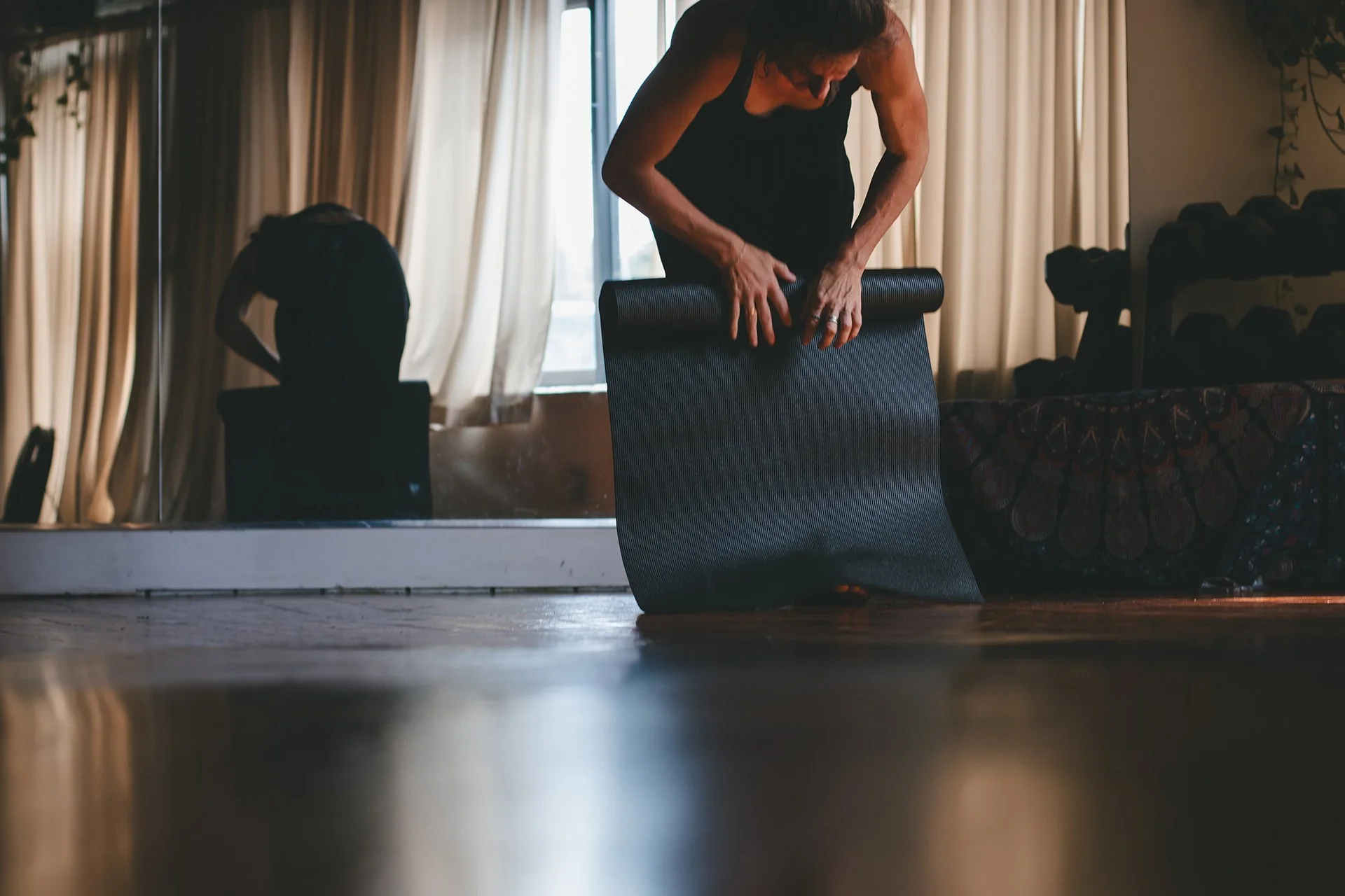 Person bending over and rolling out a yoga mat in a room with large windows and curtains.