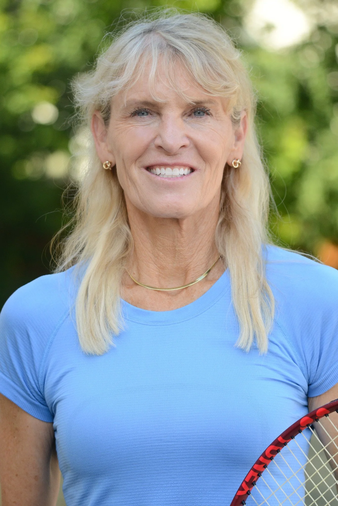 A smiling older woman with blond hair, wearing a light blue athletic shirt, gold earrings, a gold necklace, and holding a tennis racket outdoors with a blurred green background.