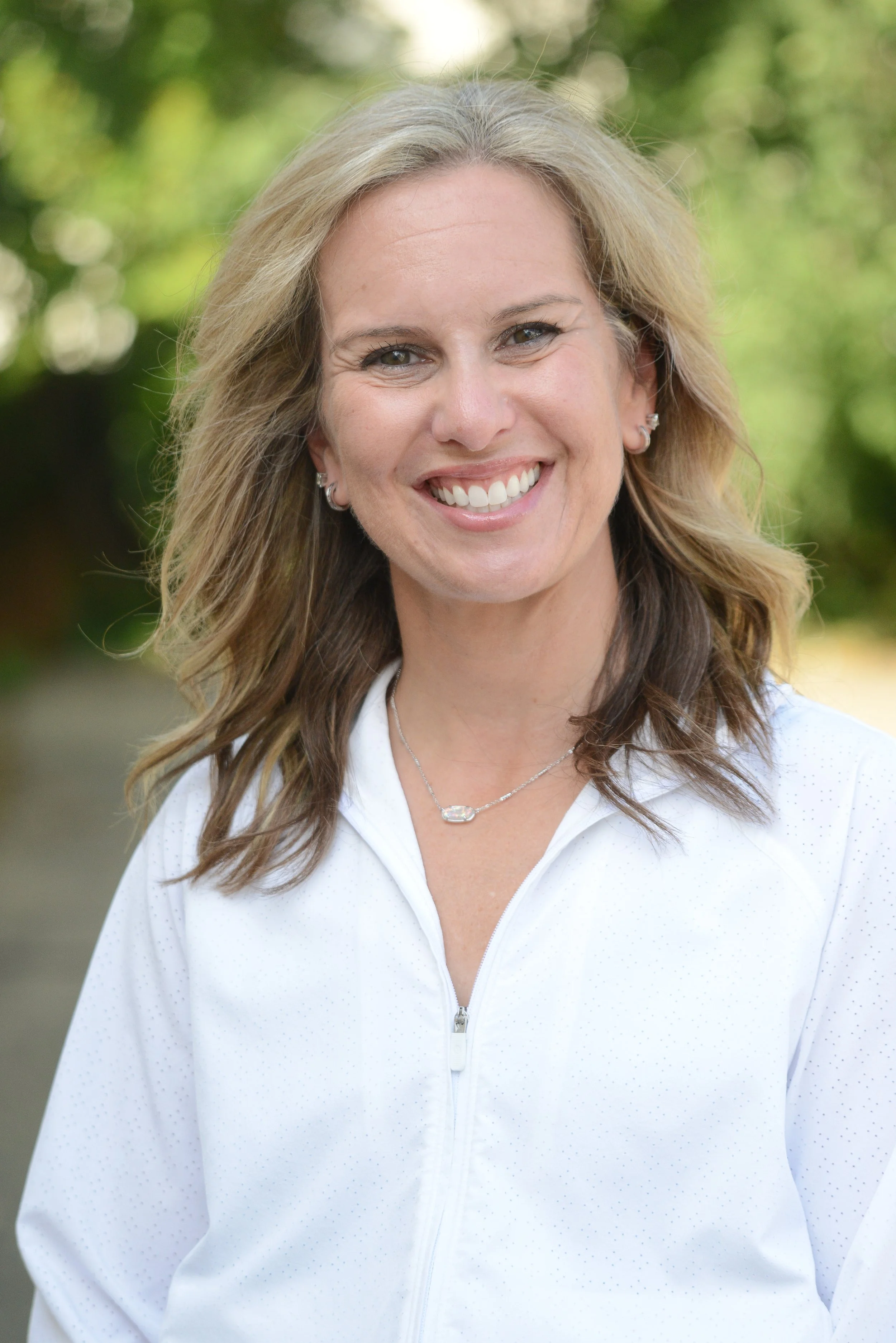Smiling woman outdoors with green foliage background, wearing a white zip-up jacket, earrings, and a necklace.
