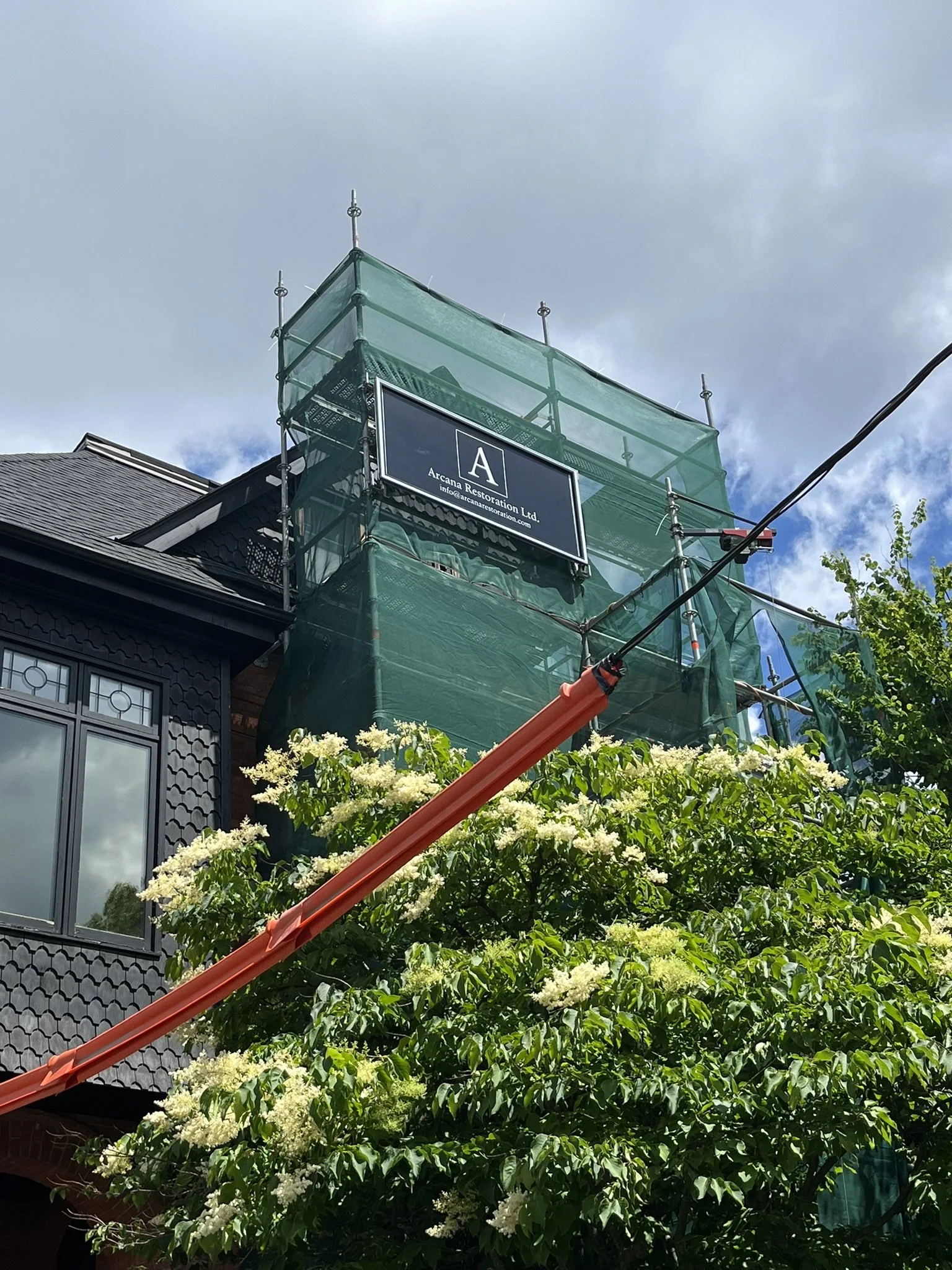 Green dust netting, Arcana Restoration sign, power line coverings at the front of a heritage house with flowering tree in front