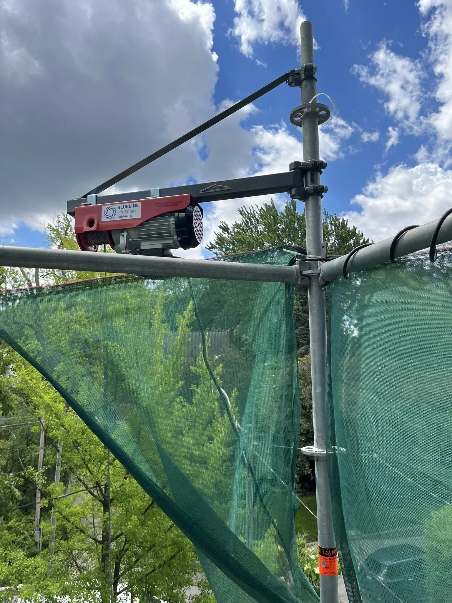 Electric materials hoist on scaffold with dust netting above trees