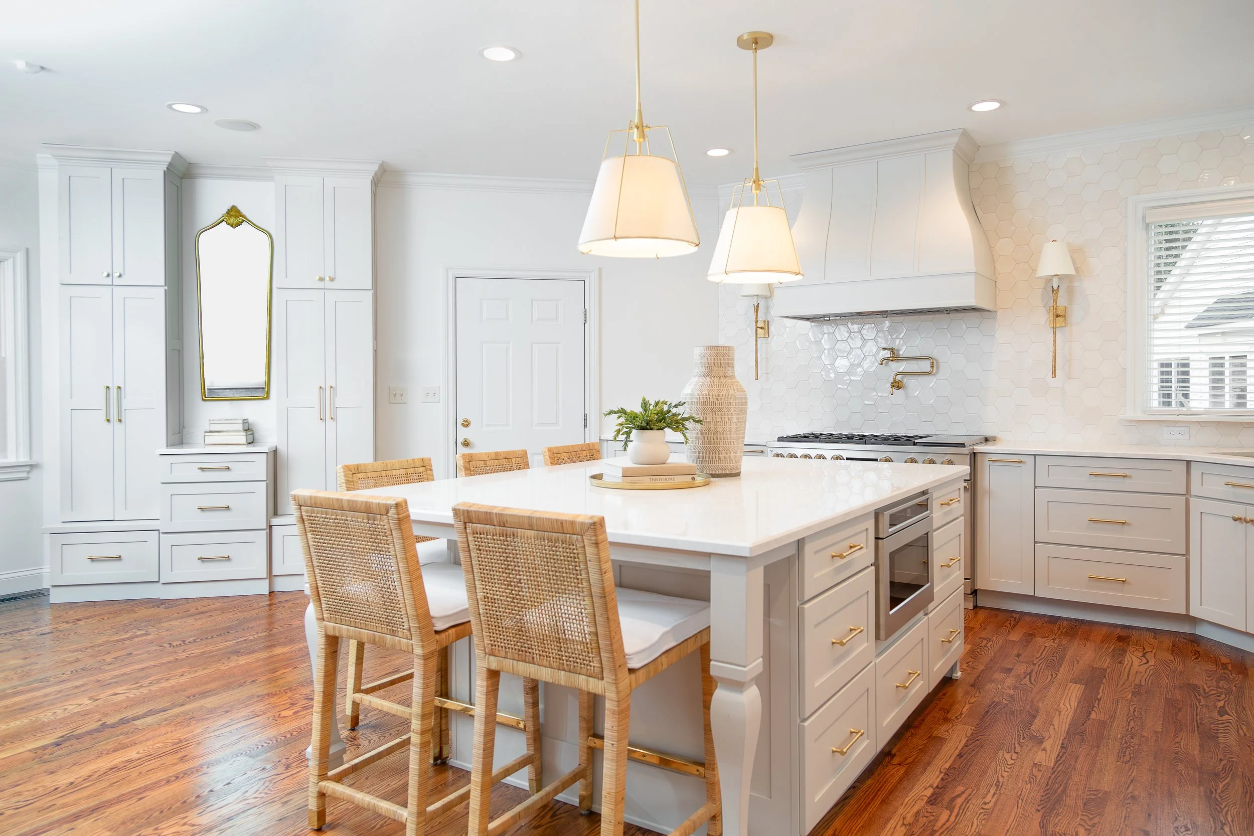 Modern kitchen with white cabinetry, gold hardware, a white island, and wicker chairs. Pendant lights and natural light from a window.  custom cabinets, Missouri cabinet maker. traditional kitchen, classic