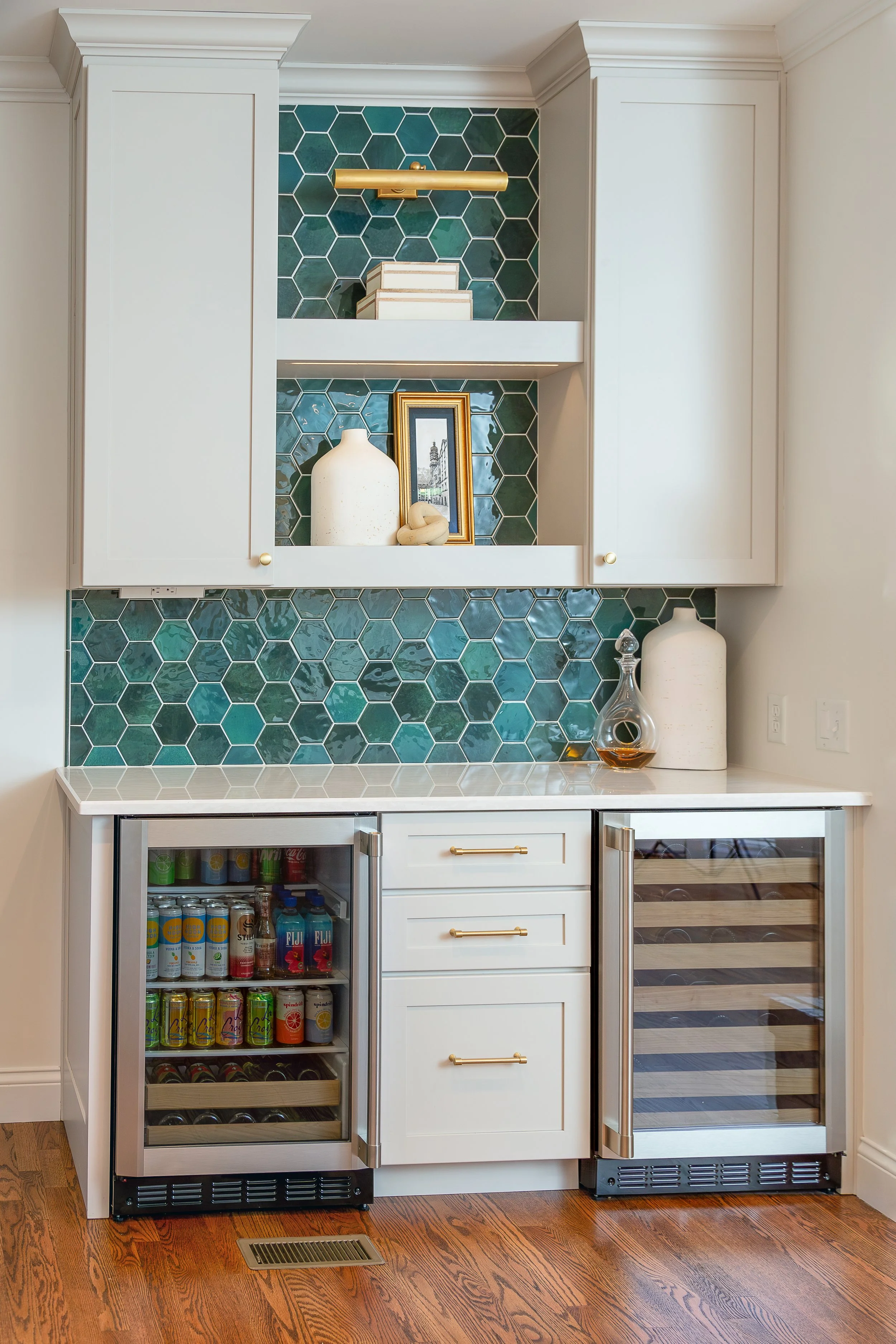 A modern kitchen with white cabinetry, teal hexagonal tile backsplash, and a built-in wine refrigerator showing beverages inside. Mini bar, coffee station custom cabinets, Missouri cabinet maker.