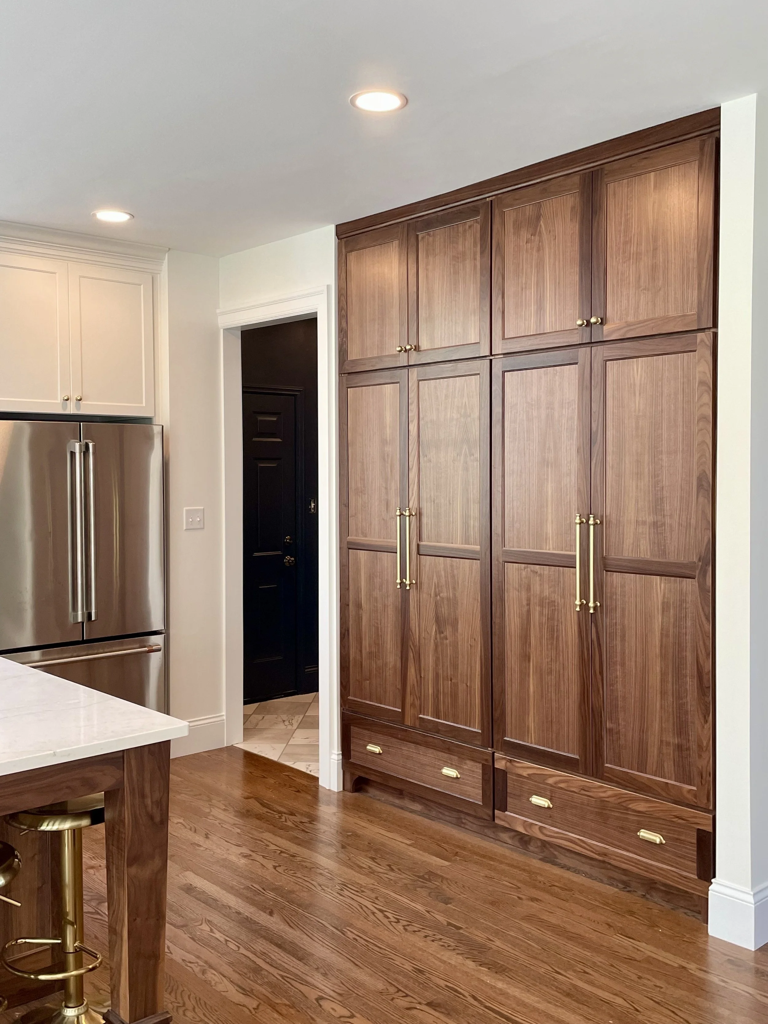 Kitchen interior with dark wooden cabinets, a stainless steel refrigerator, hardwood floor, and a small portion of a white kitchen island with a wooden leg.  large pantry custom cabinets, Missouri cabinet maker.