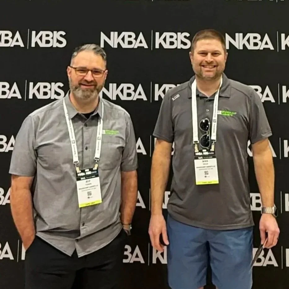 Two men standing in front of a black backdrop with white logos that say 'NKBA' and 'KBS'. Both are wearing grey shirts with conference badges hanging around their necks and are smiling.