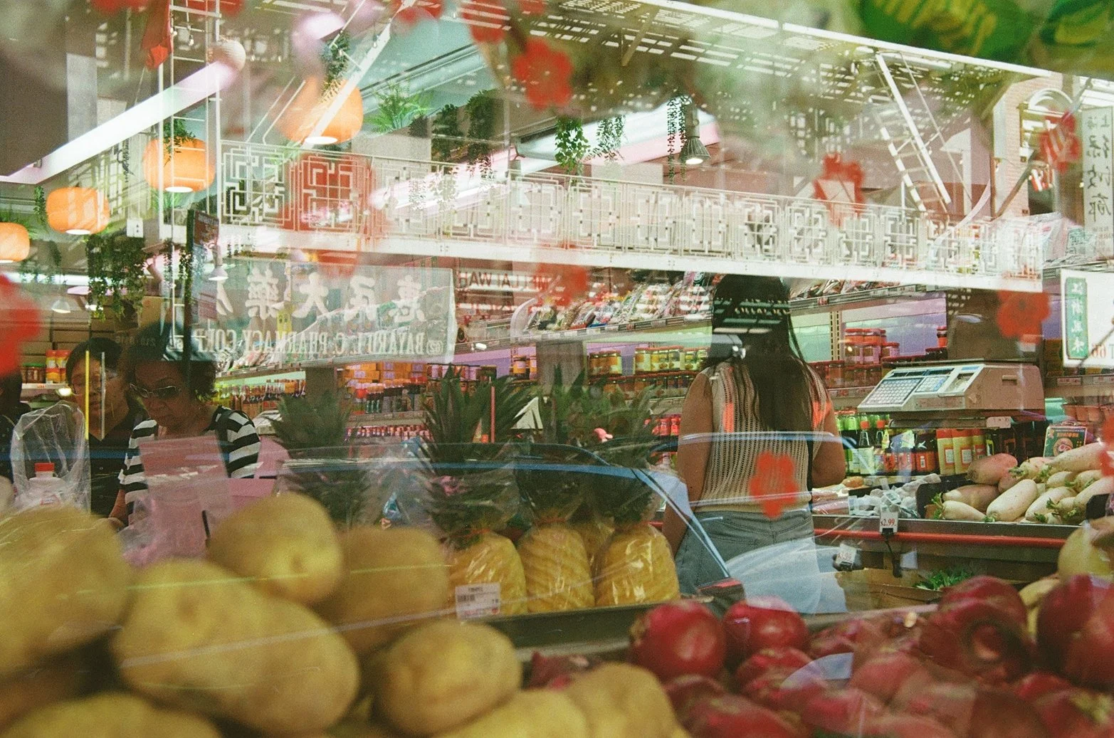 Vista de un supermercado asiático desde afuera, con frutas y verduras en el mostrador y personas comprando, reflejado en la ventana con decoraciones y letreros en chino.