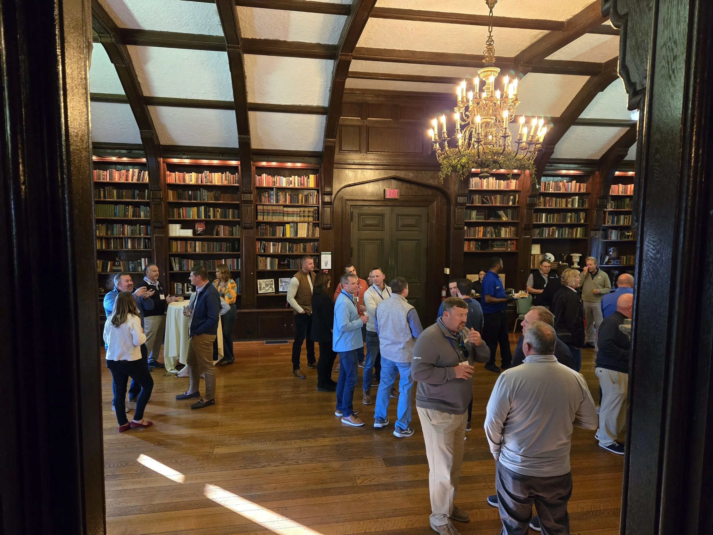 People socializing in a large, elegant library room with wooden bookcases and a chandelier.