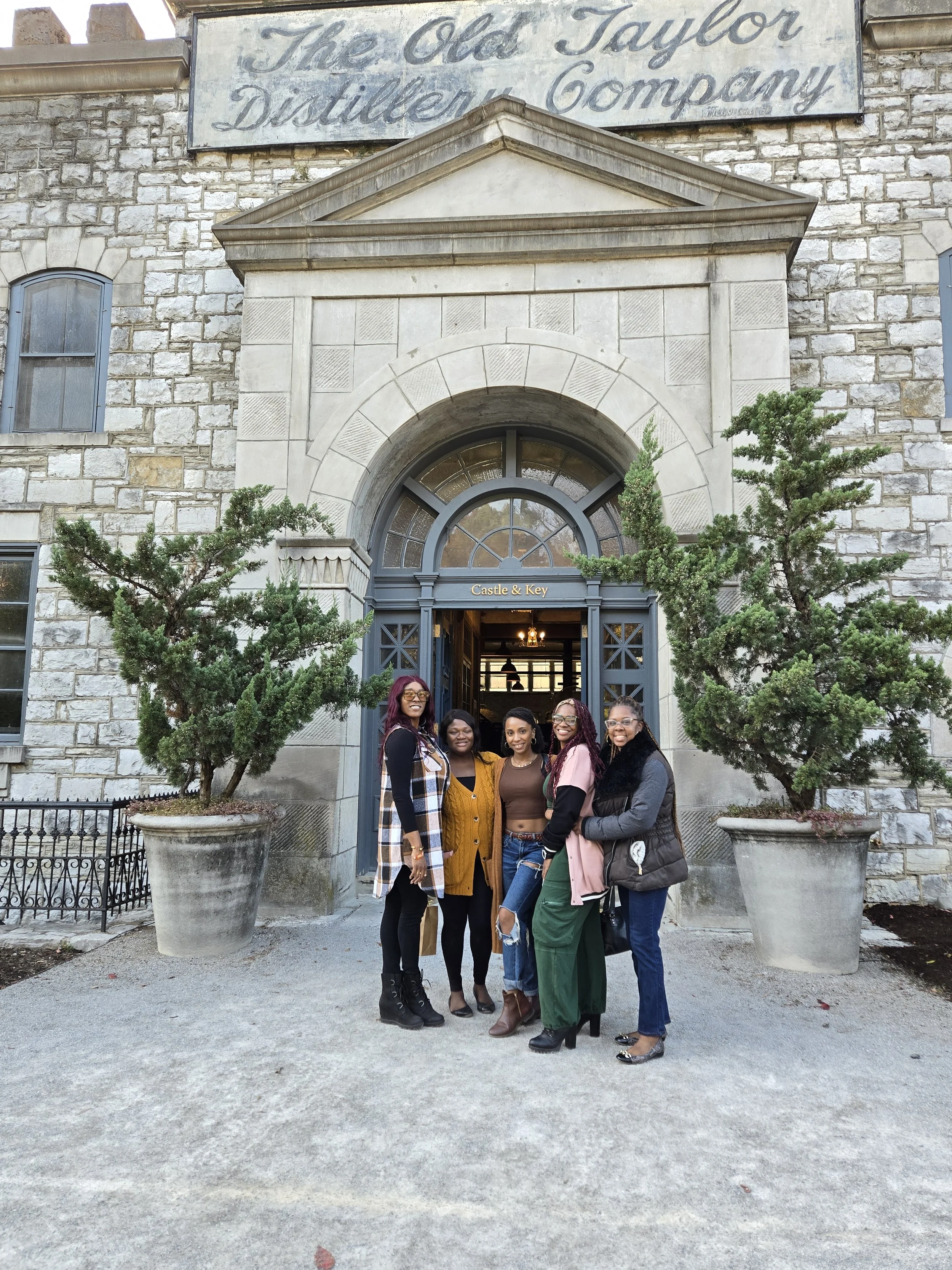 Five women standing together in front of the entrance to The Old Taylor Distillery Company, smiling. The entrance has a large arched doorway with a sign above that reads "Castle & Key". There are two large potted trees on either side of the entrance