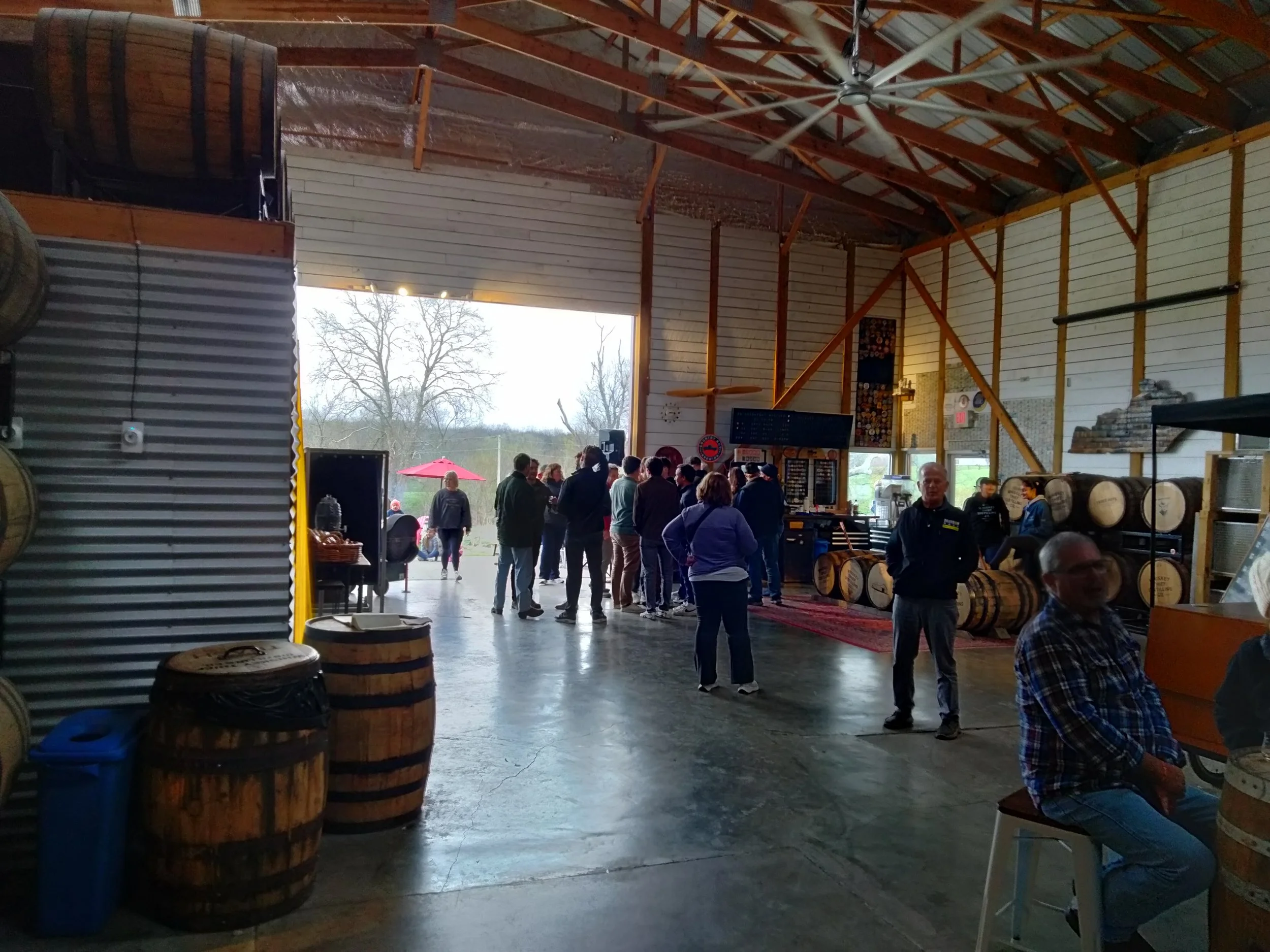 People inside a winery or brewery warehouse, some waiting in line, others talking, with wine barrels and outdoor scenery visible through the open door.