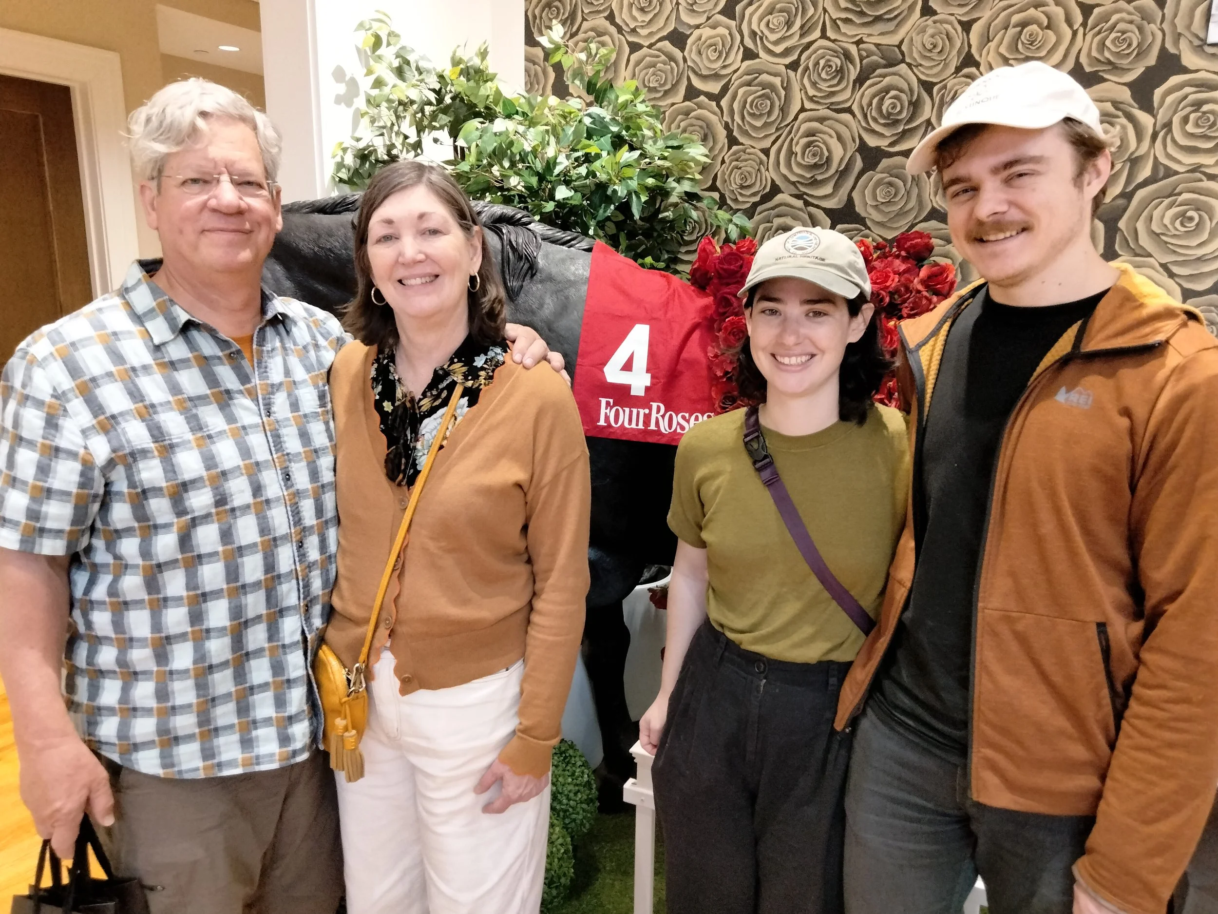 Family of four standing indoors in front of a large flower display with a red flag that has the number 4 and 'Four Roses' written on it. The group includes an older man and woman, and a younger man and woman, all smiling at the camera.