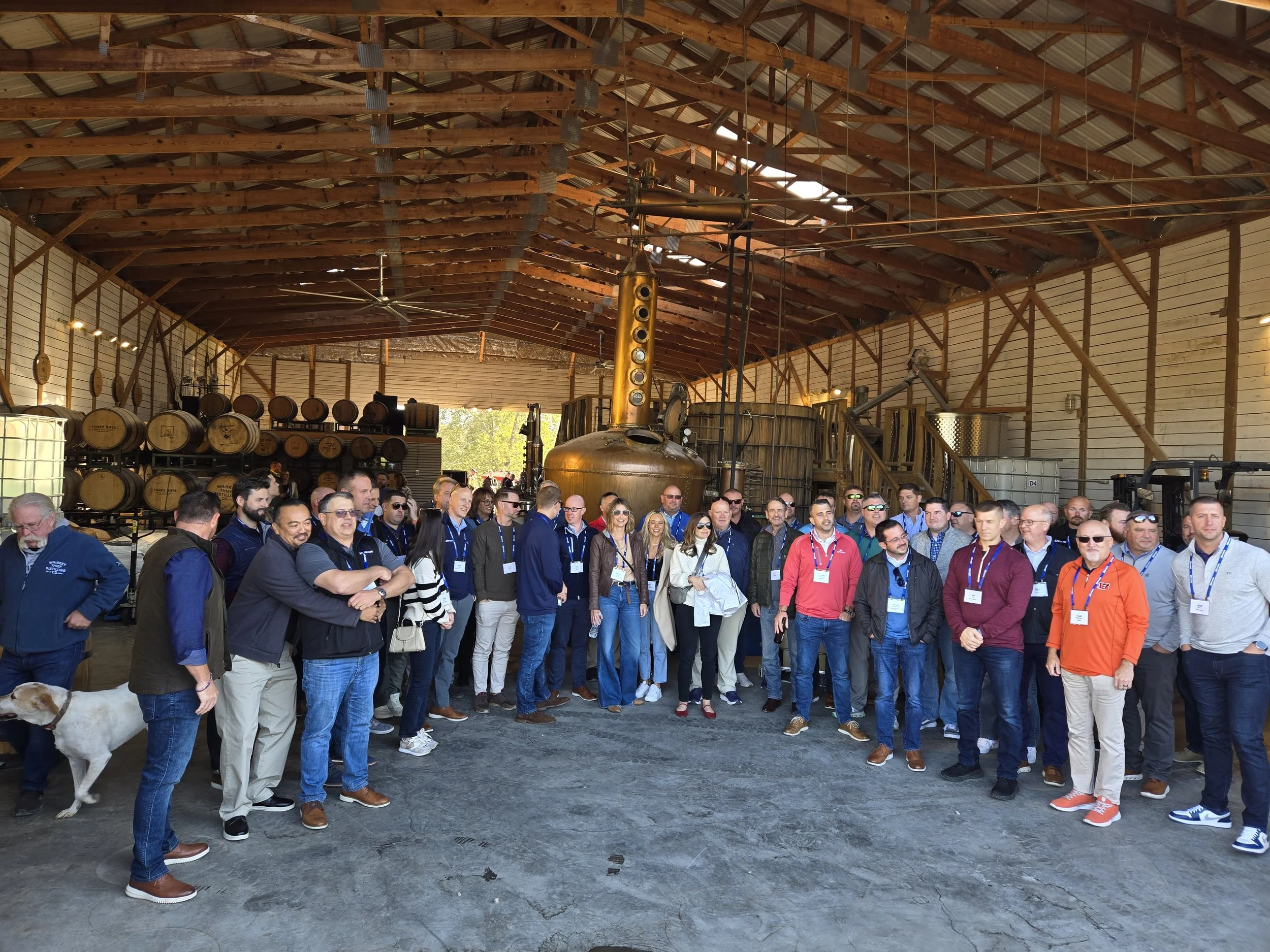 A large group of people standing together inside a rustic warehouse with wooden barrels, a copper still, and industrial equipment in the background.