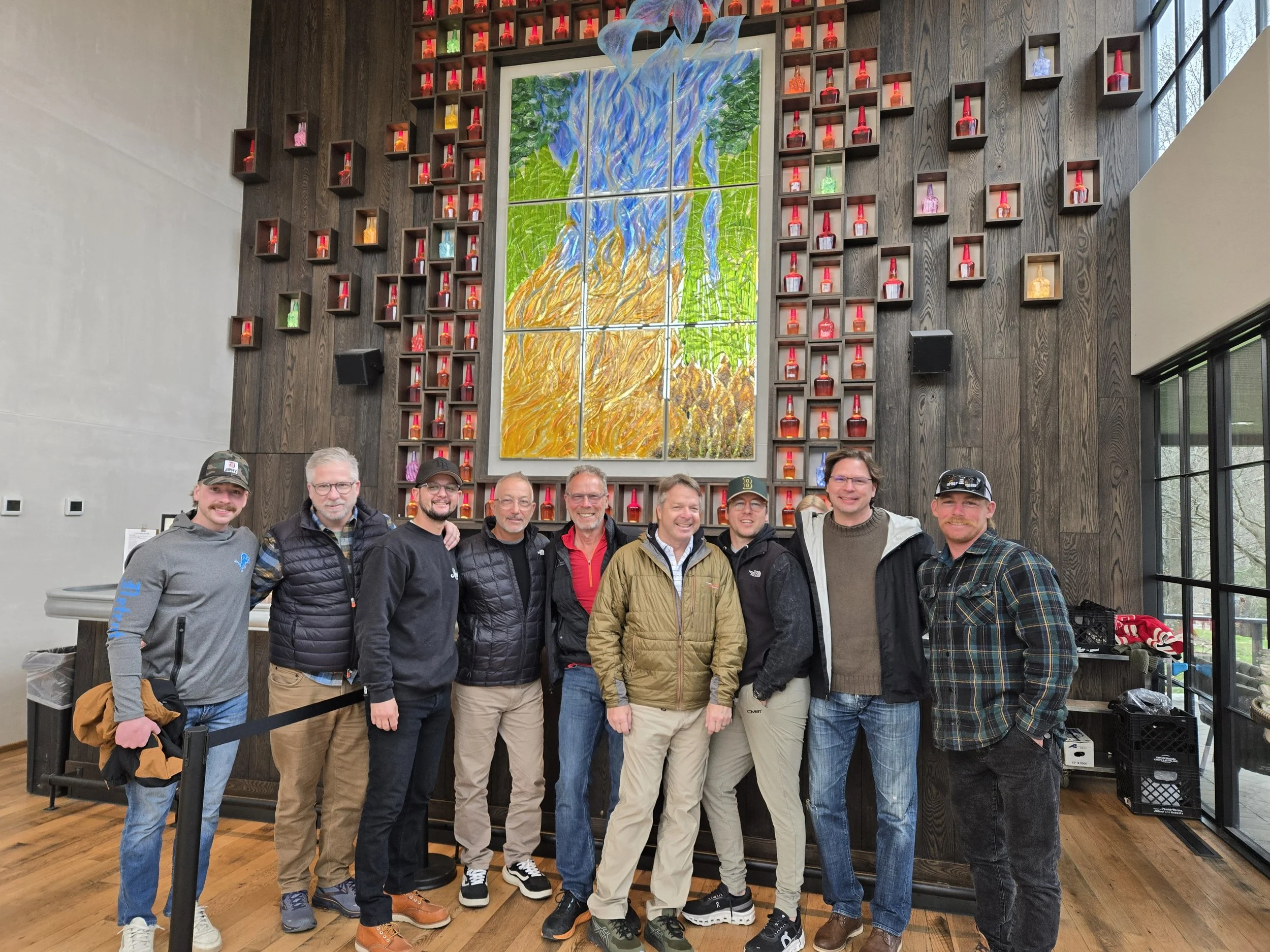 A group of nine men standing inside a building in front of a large colorful stained glass artwork depicting a tree with orange and blue leaves, surrounded by small shelves holding colorful bottles.