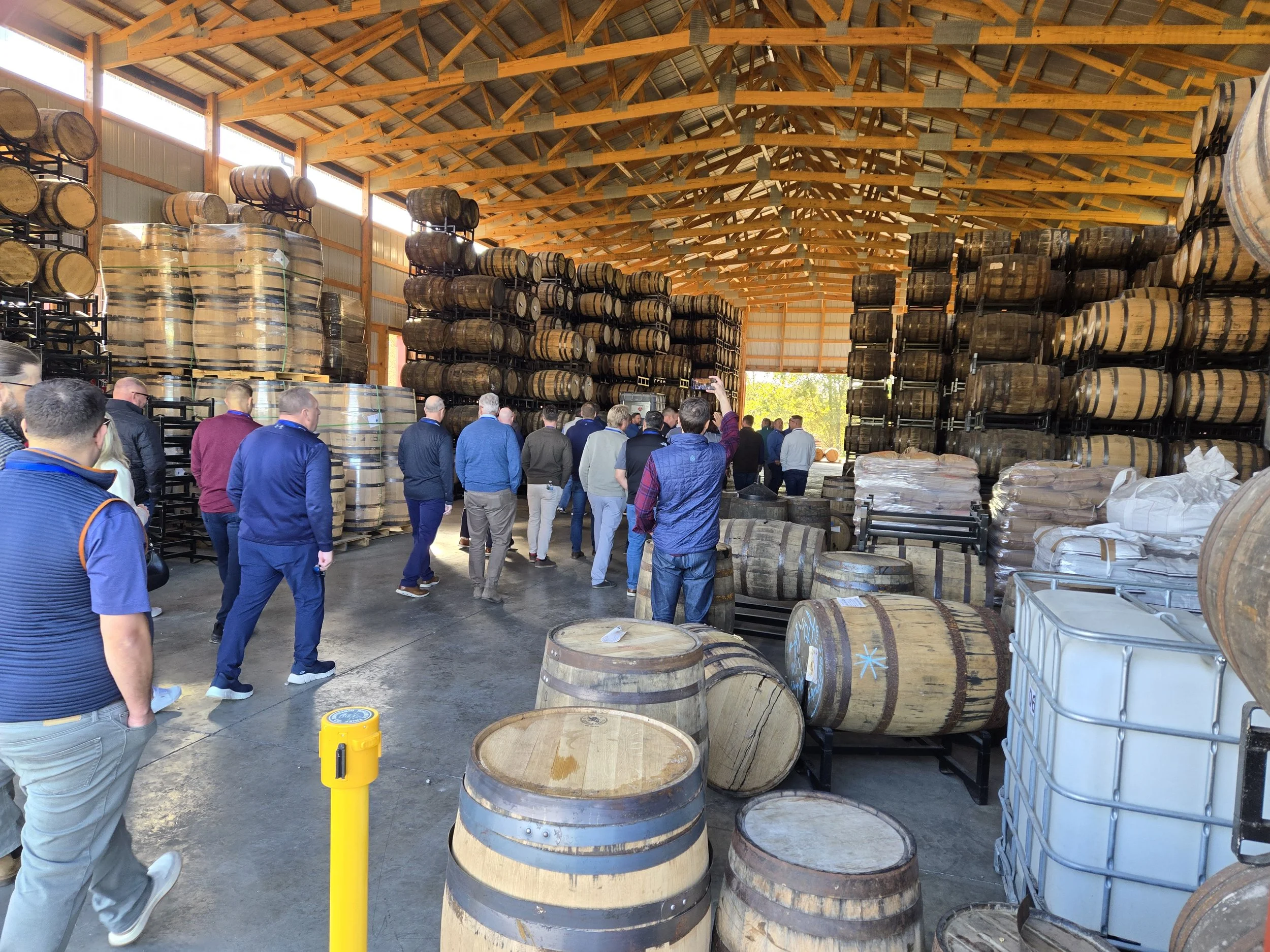 Group of people touring a large warehouse filled with stacked wooden barrels used for aging spirits.
