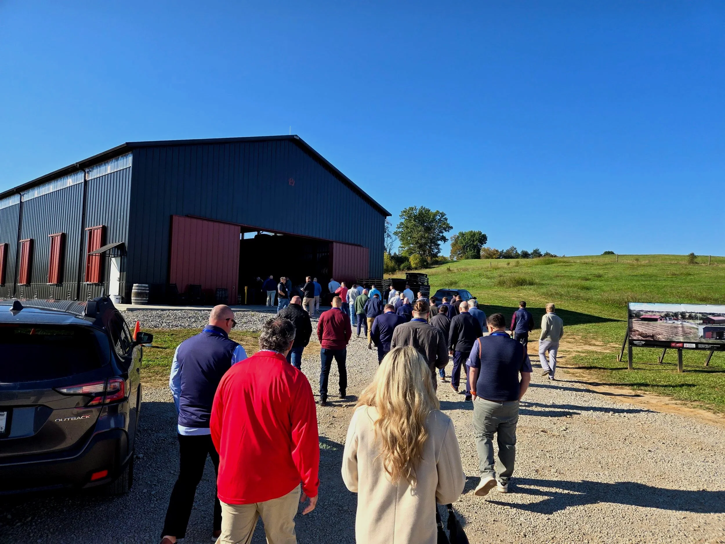People lined up outside a large dark blue barn on a sunny day with a grassy hill in the background.