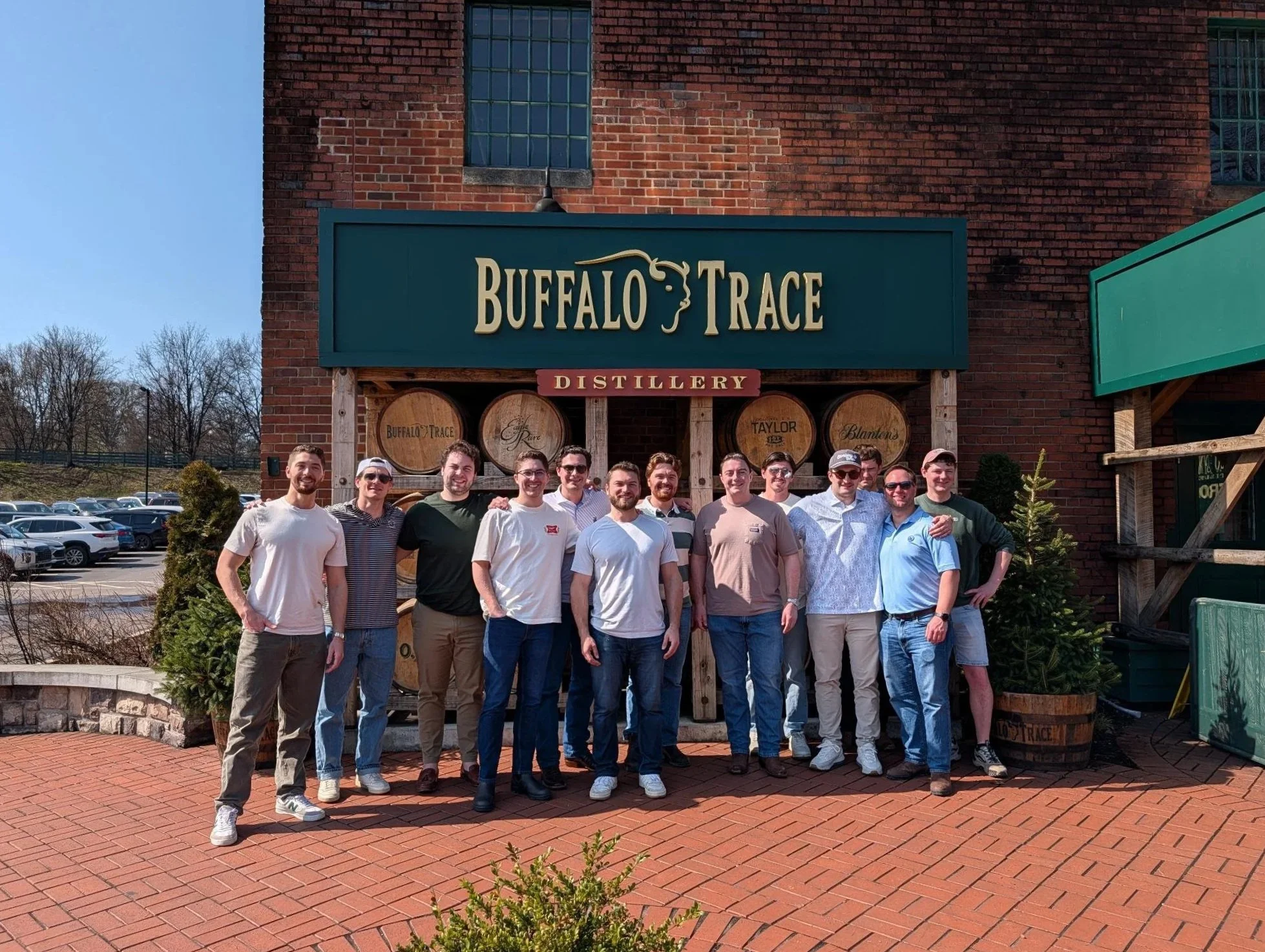 A group of people standing in front of the Buffalo Trace Distillery sign outside a brick building on a sunny day.