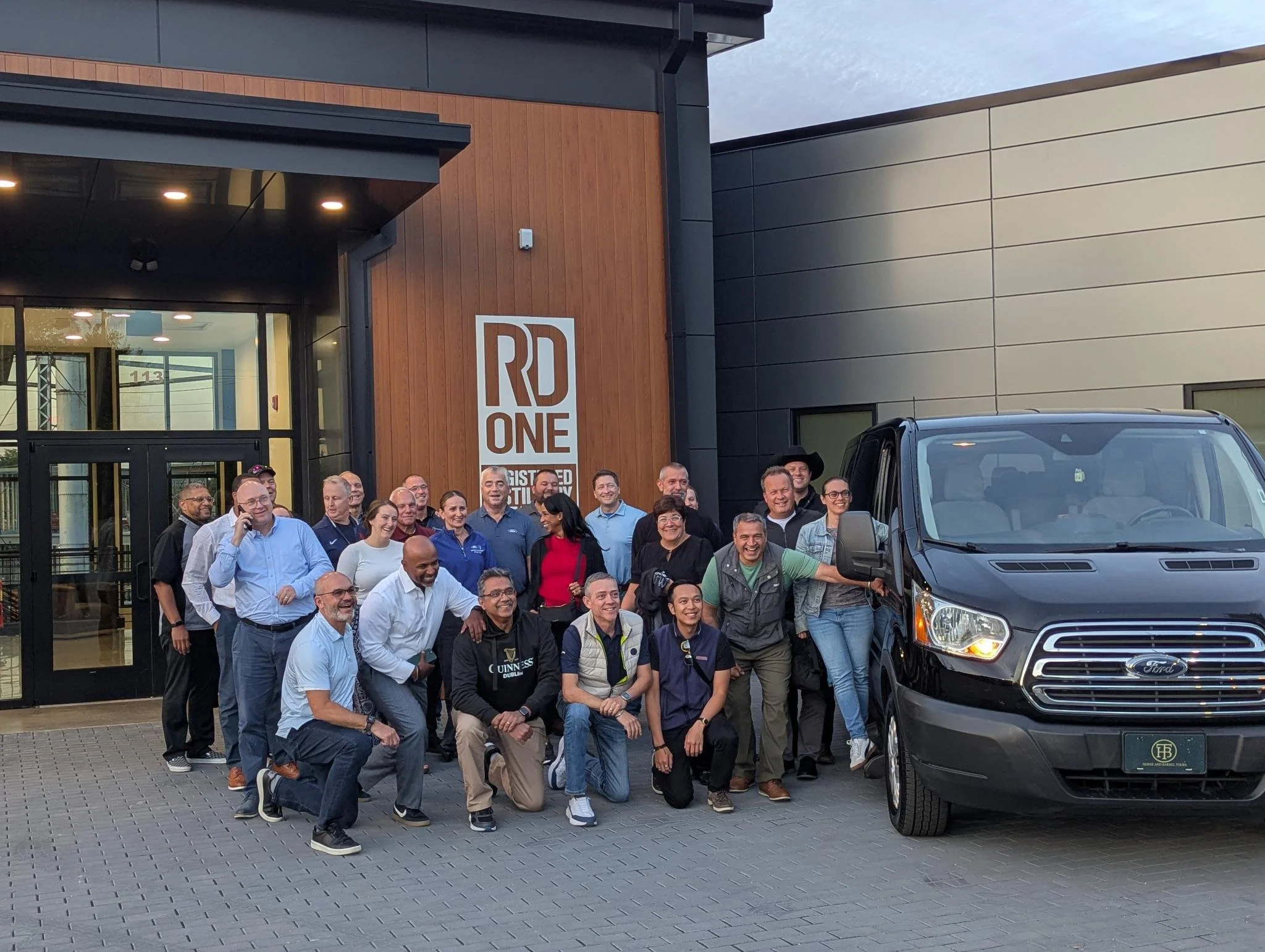 A group of people gathered outside a modern building with wood and metal paneling. They are smiling and posing for the photo in front of a black vehicle. The building has a large sign that says 'RD ONE' and large windows. The sky is partly cloudy.