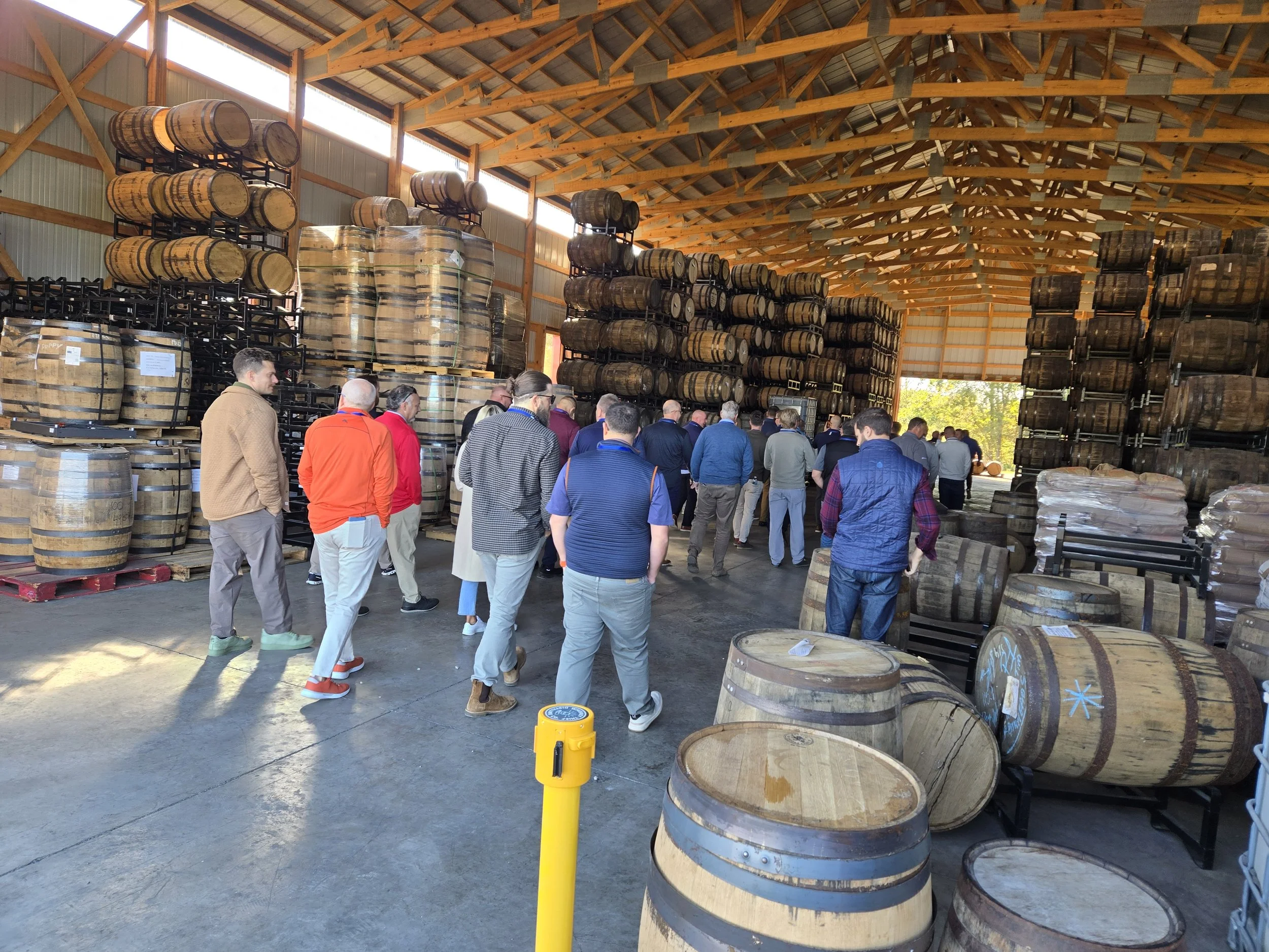 People touring a warehouse filled with stacked barrels used for aging whiskey or other spirits, with some barrels placed on racks and others on the floor.