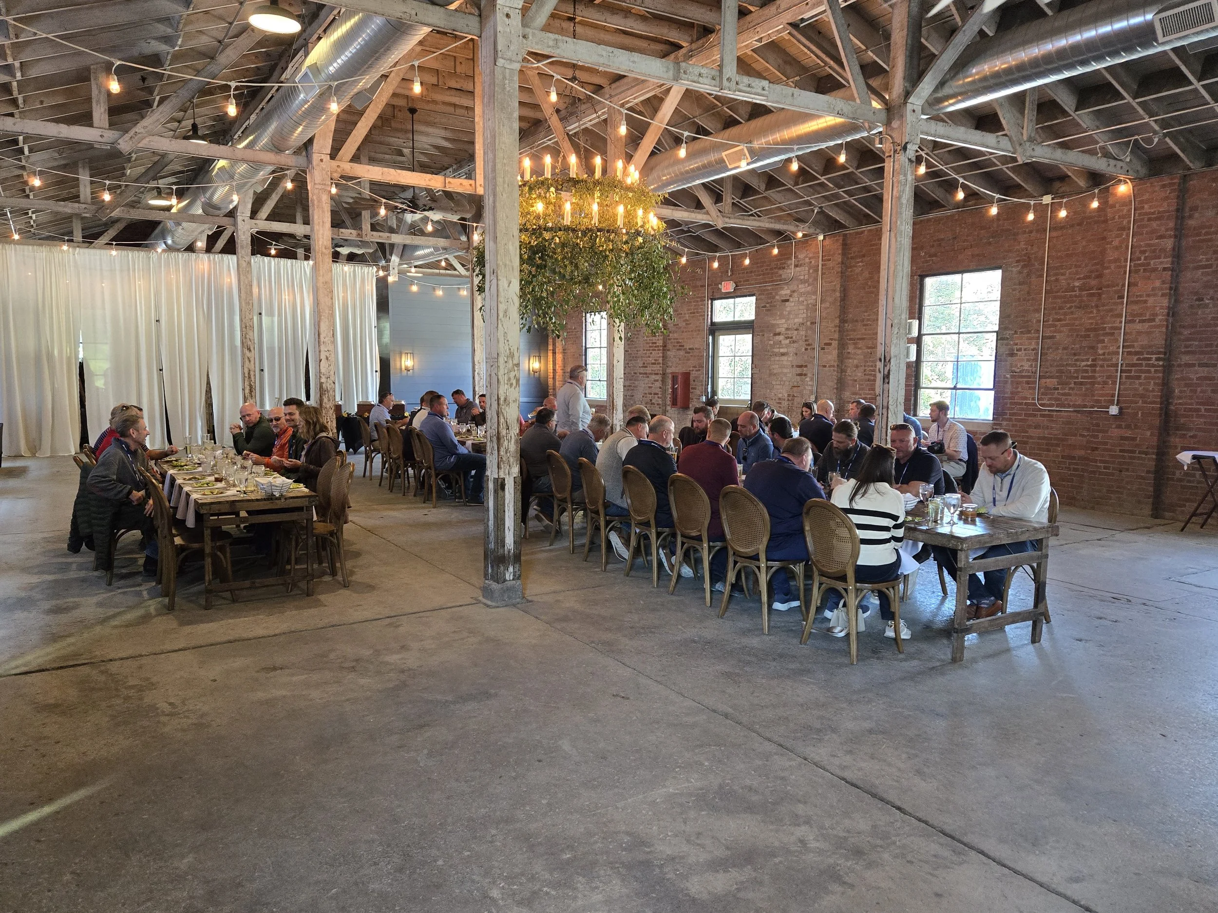 People seated at long tables dining in a large, rustic warehouse with exposed brick walls, wooden beams, string lights, and large windows.